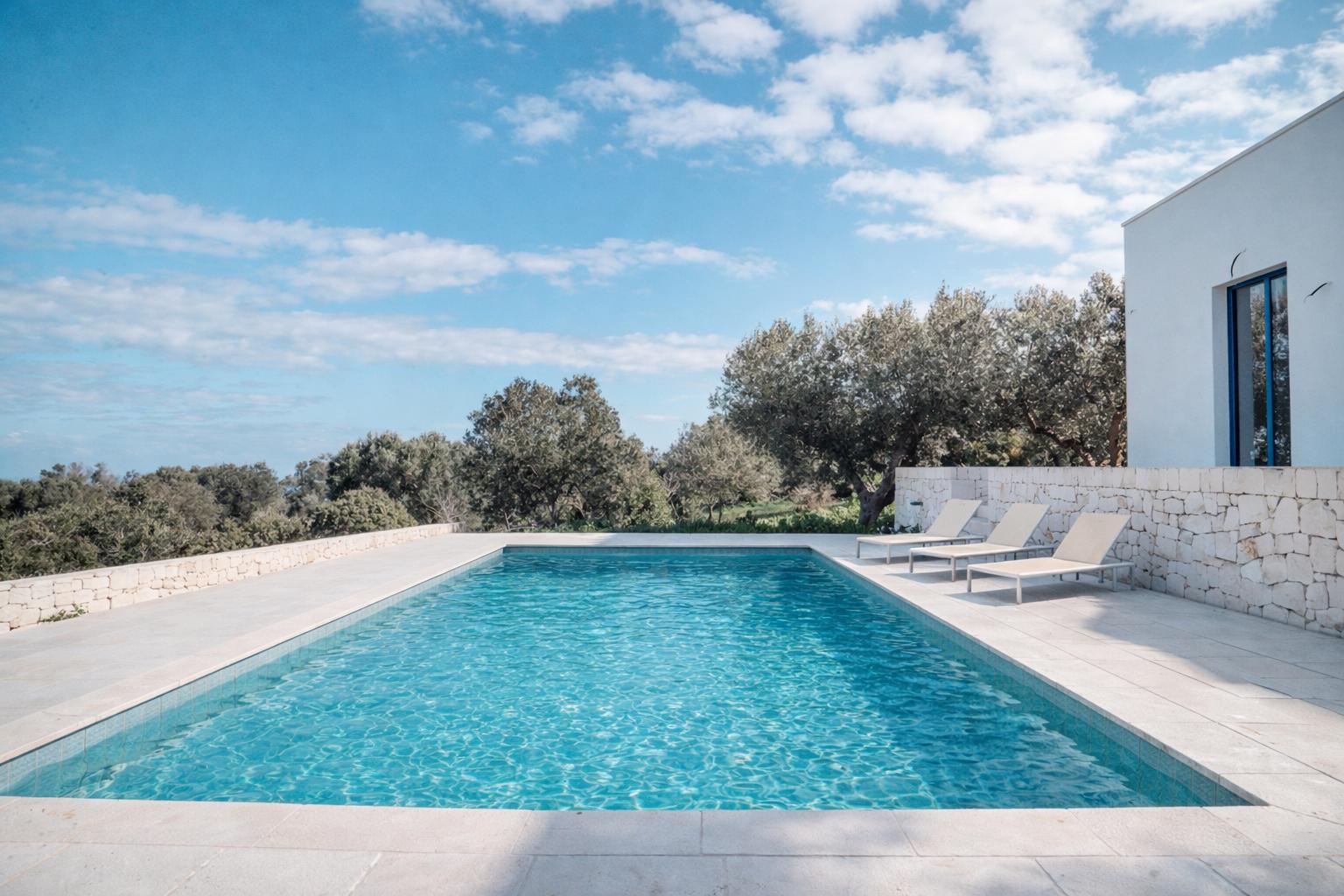 Pool with loungers, stone wall, and olive trees under blue sky.
