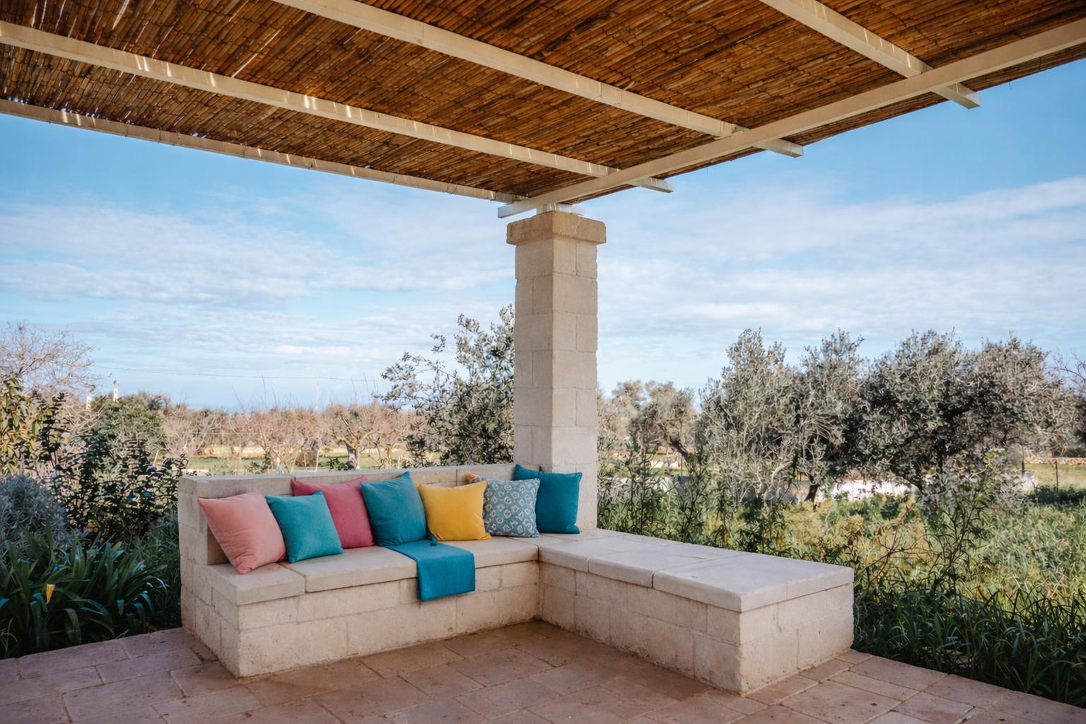 Terrasse mit Steinbank, bunten Kissen und strohgedecktem Dach. Blick auf Olivenhaine.