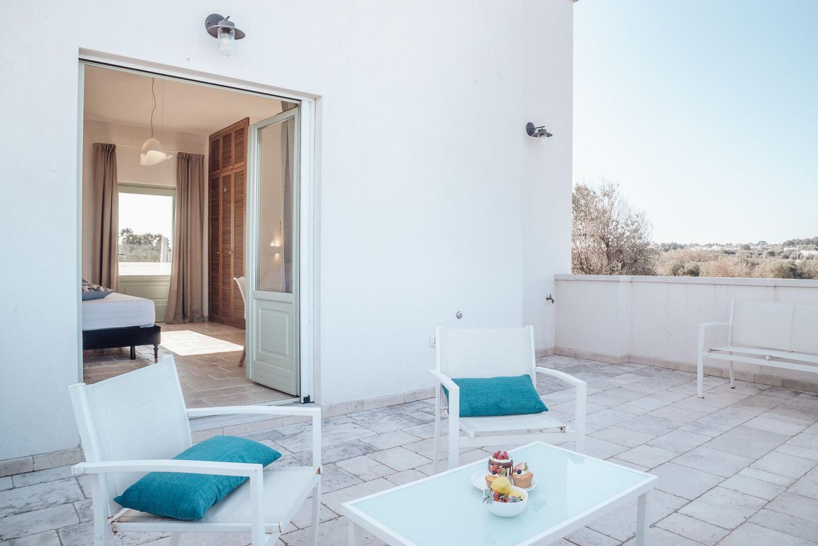 Terrasse mit weißen Stühlen und Tisch. Blick in Schlafzimmer mit Bett und Vorhängen.