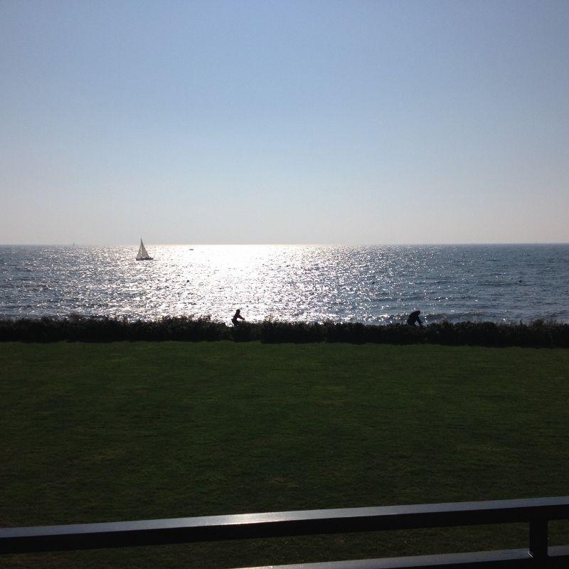 Meer mit Segelboot und zwei Personen im Gras.