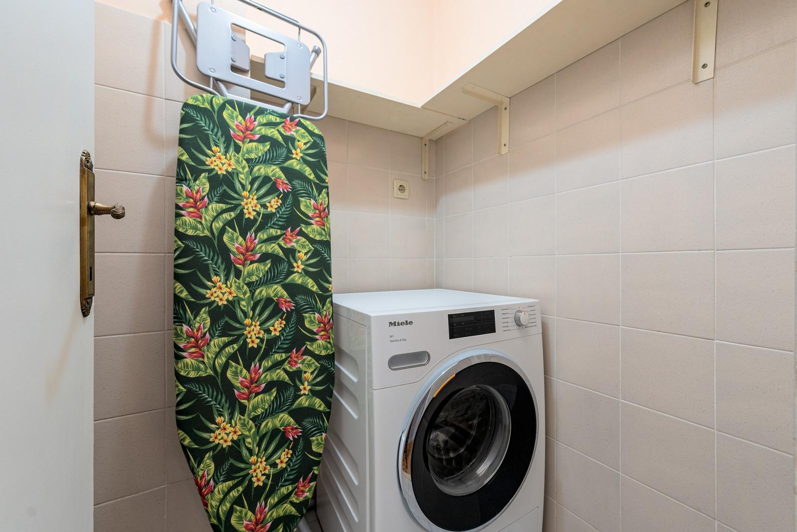 Waschmaschine und Bügelbrett mit tropischem Muster in einem Raum mit Fliesenwänden.