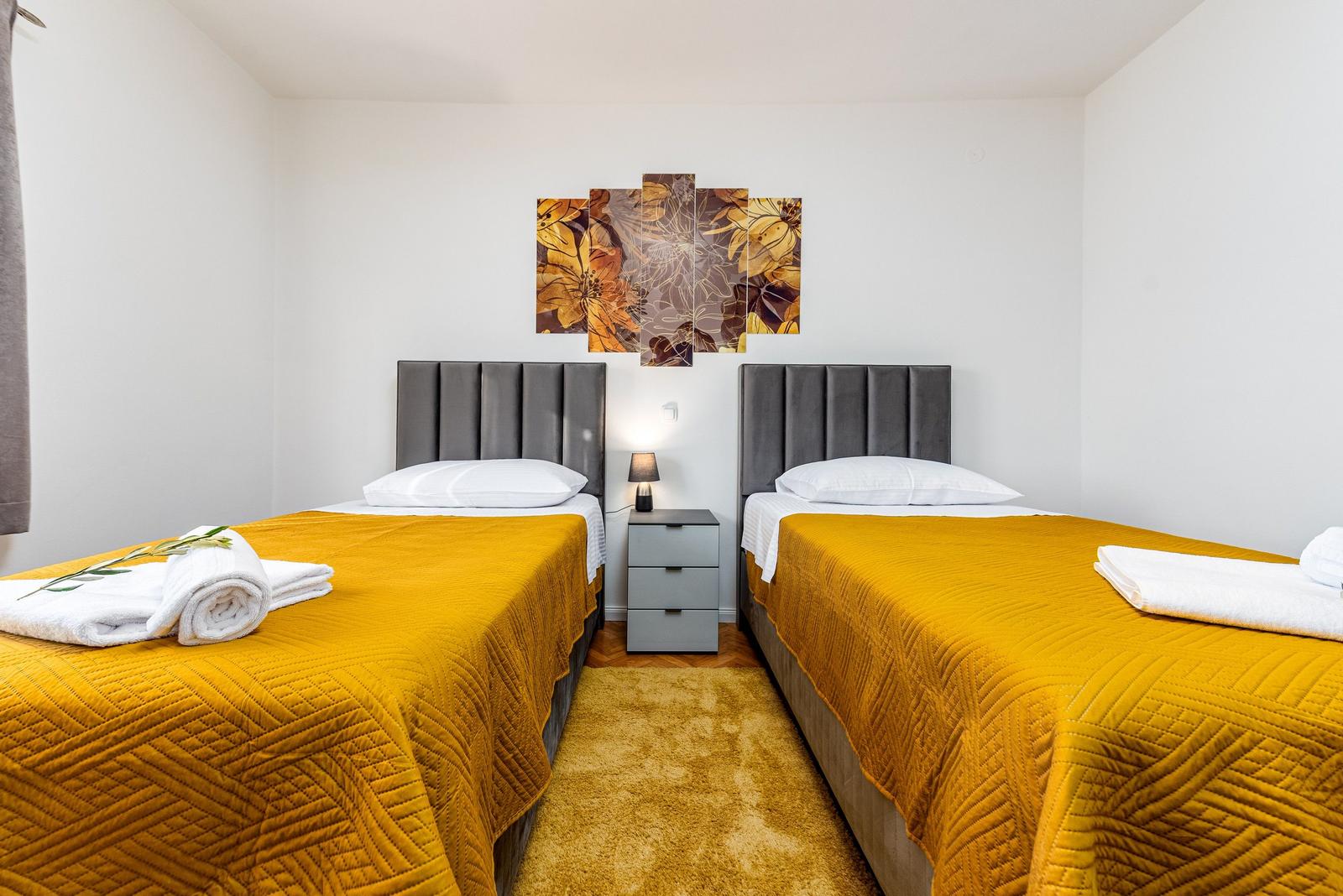 Zwei Betten mit gelben Bezügen und grauen Kopfenden. Zwischen ihnen steht ein Nachttisch mit Lampe.