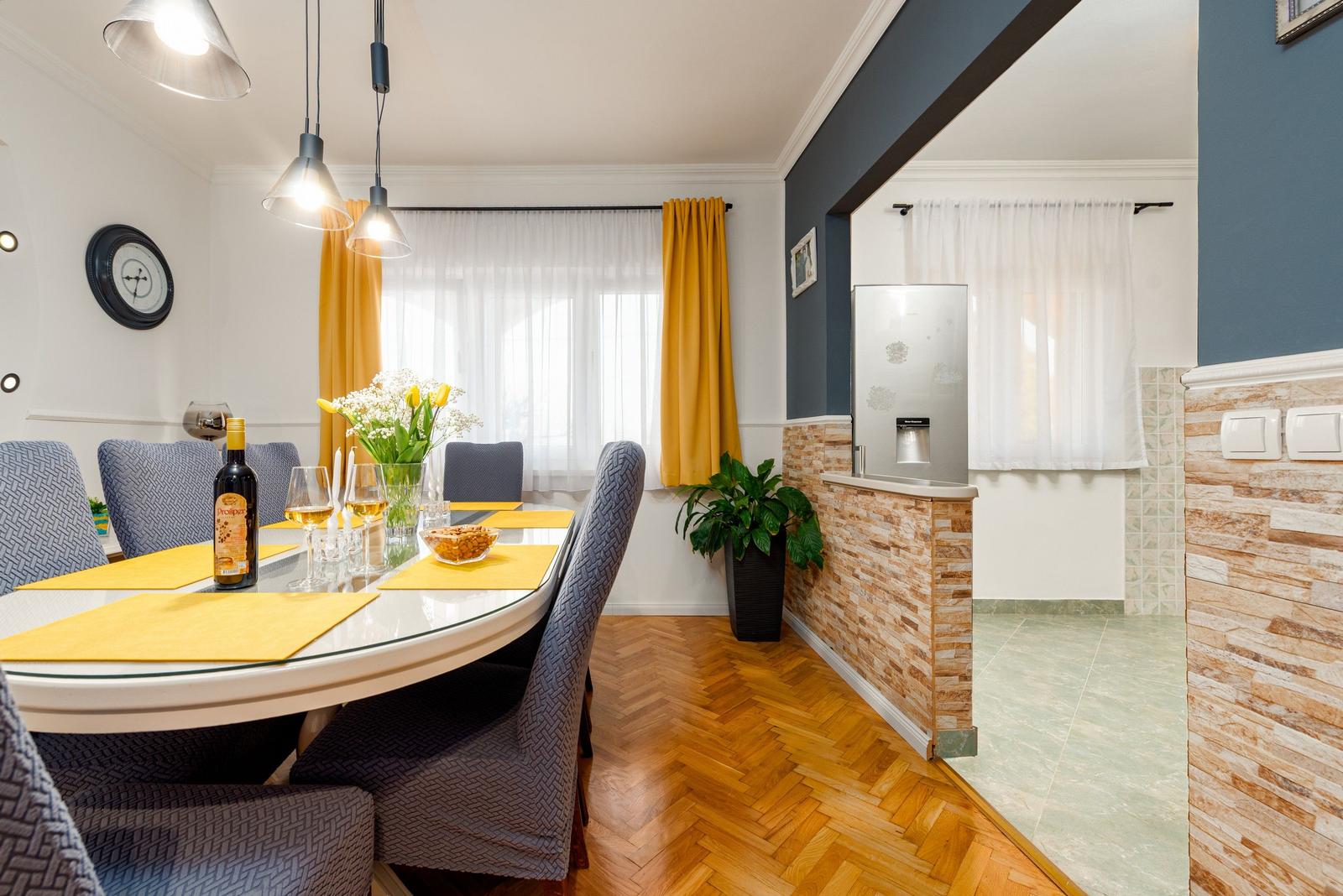 Essbereich mit runder Tisch, gelben Tischdecken und grauen Stühlen. Hintergrund: Küche mit Kühlschrank und Steinwand.