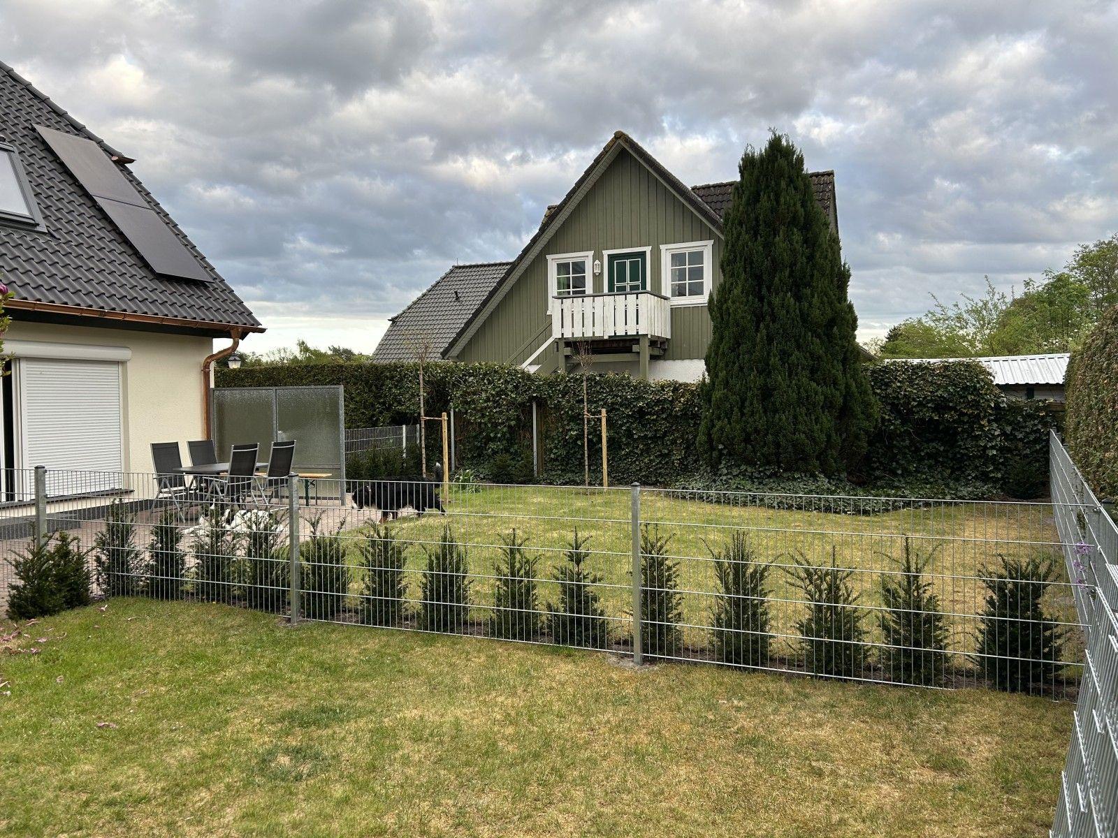 Haus mit Garten, Terrasse und Holzveranda. Umzäunt mit Metallzaun und Beerensträuchern.