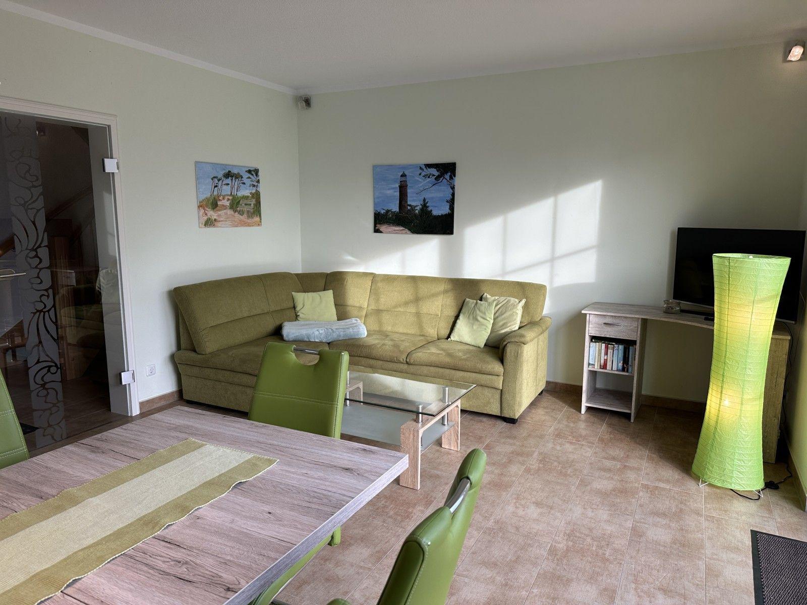 Wohn- und Essbereich mit grünem Sofa, Tisch und Stühlen sowie Fernseher und Stehlampe.