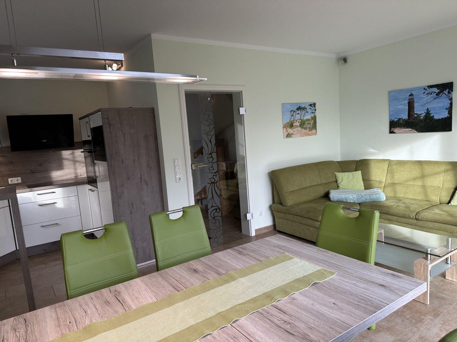 Wohn- und Essbereich mit grüner Couch und Tischstühlen. Küche mit TV und Küchenschränken.