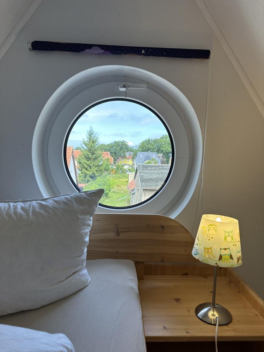 Schlafzimmer mit runden Fenster und Holzbett. Lampe und Blick auf Außenbereich.