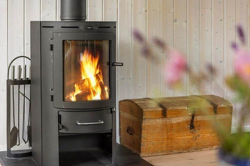 Wood stove with burning logs, next to wooden chest and flowers in foreground.