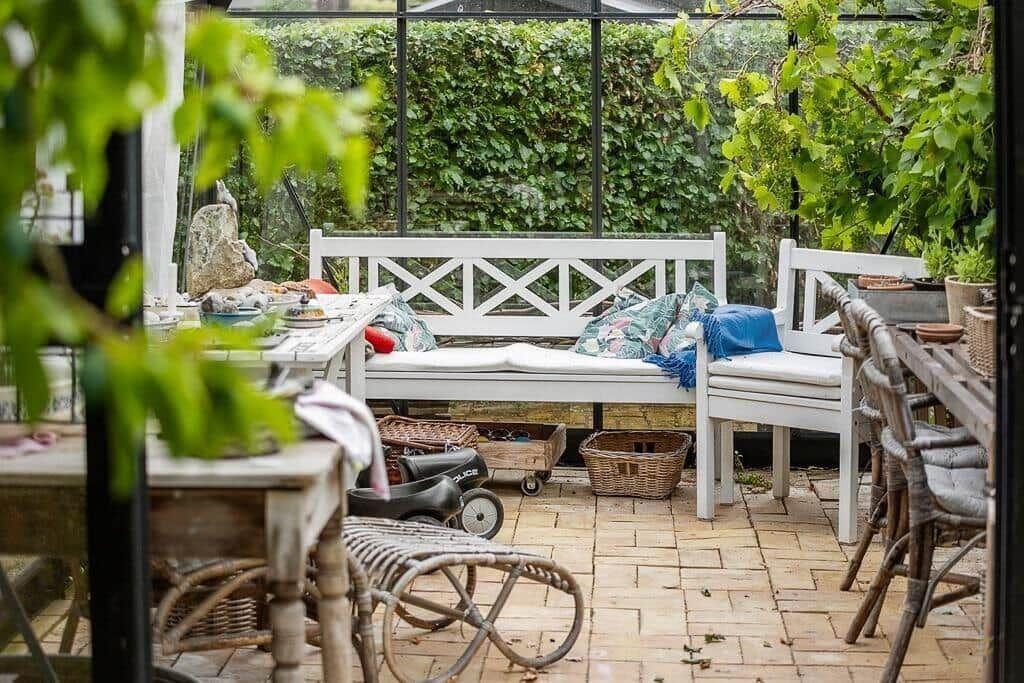 Terrace with white sofa, tables and chairs. Background: green hedge and glass wall.