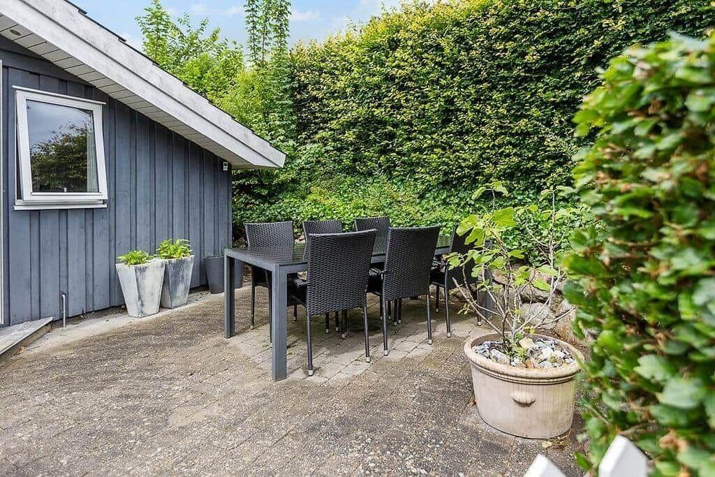 Terrace with table and chairs in front of gray house with hedge.