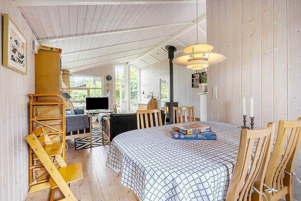Dining room with wooden table, chairs, and tablecloth. Background shows living area with fireplace and windows.
