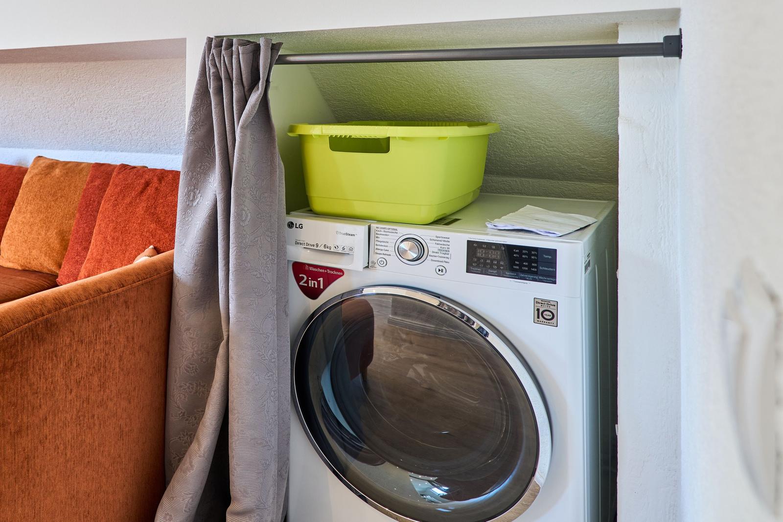 Waschmaschine mit Trocknerfunktion neben einem Sofa mit Kissen.