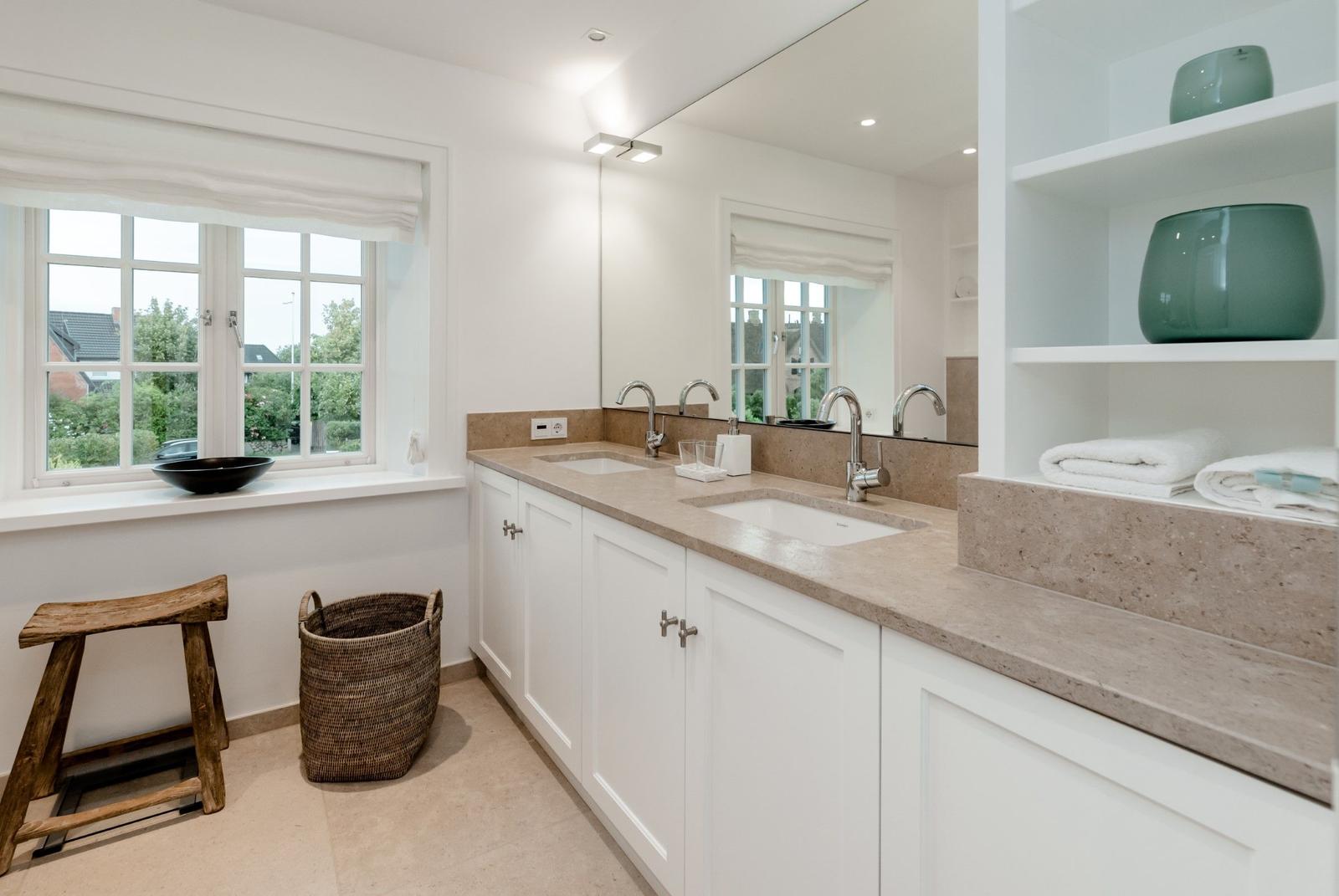 Badezimmer mit zwei Waschbecken, Spiegeln und Fenster. Holzstuhl und Korb sind vorhanden.