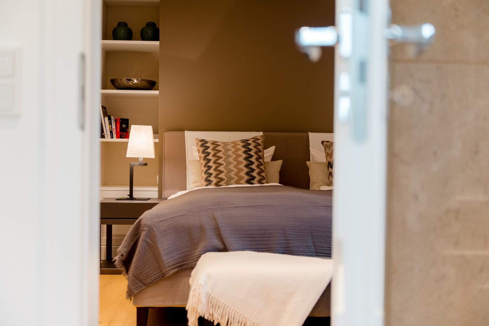 Schlafzimmer mit Bett, Kissen und Nachttisch. Hintergrundregal mit Büchern und Dekorationsgegenständen.