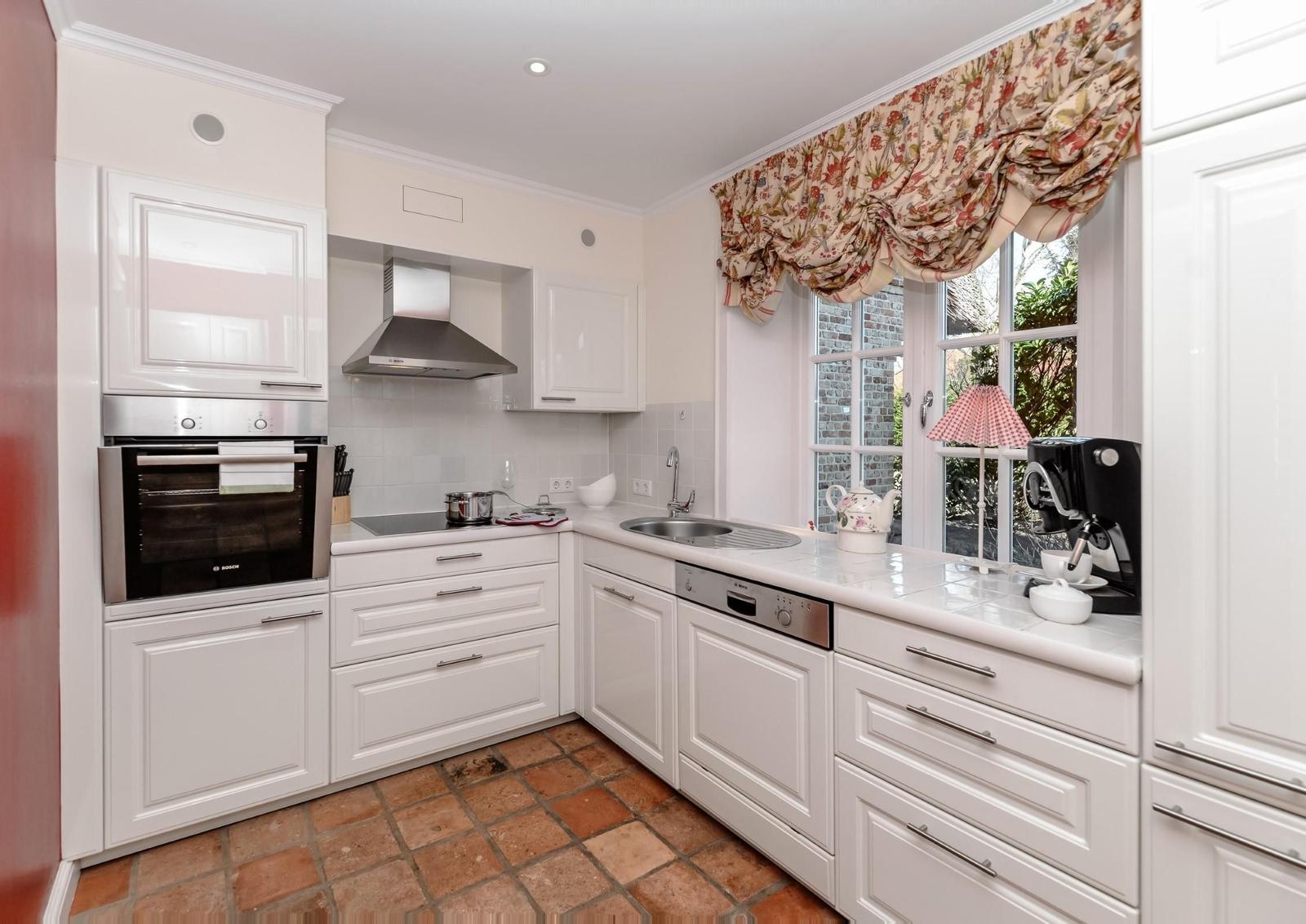 Küche mit weißen Schränken, Herd, Spüle und Fenster mit Vorhang.