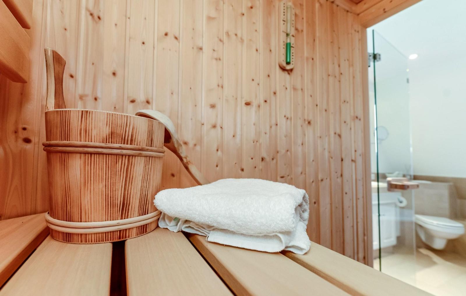 Sauna mit Holzbank, Eimer und Handtuch. Im Hintergrund Bad mit Dusche.