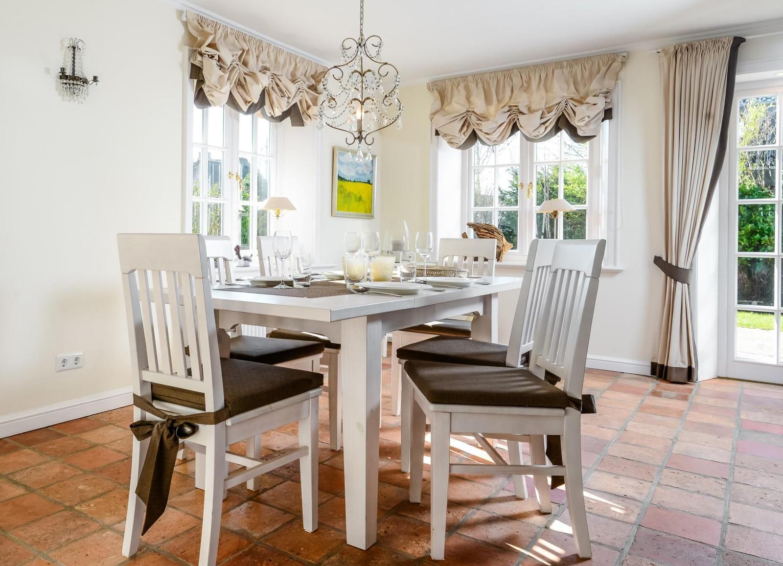 Esszimmer mit weißem Tisch, Stühlen und Terrakottboden. Fenster mit Vorhängen und Kronleuchter.