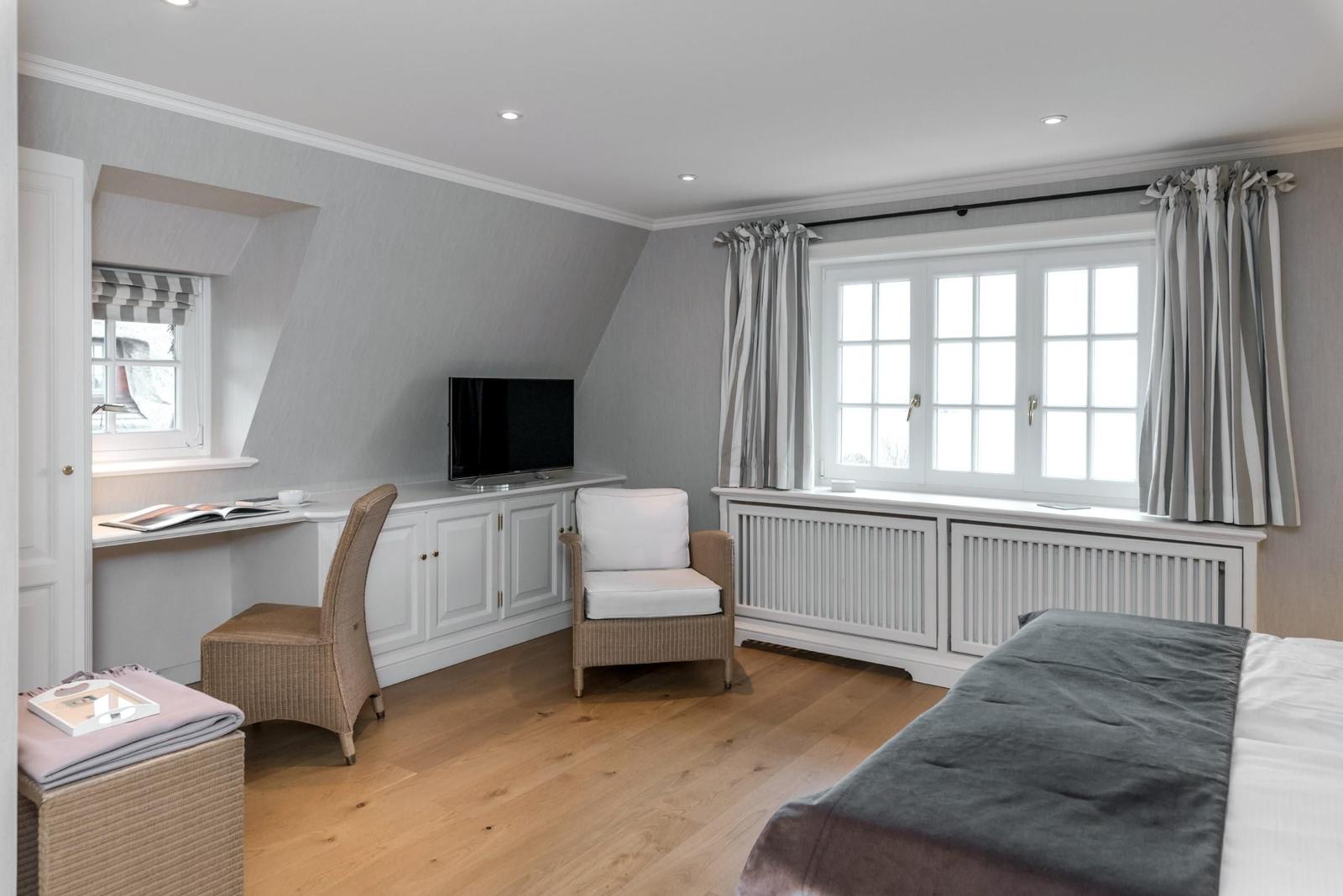 Schlafzimmer mit Bett, Schreibtisch, Sessel und TV. Holzboden und große Fenster.