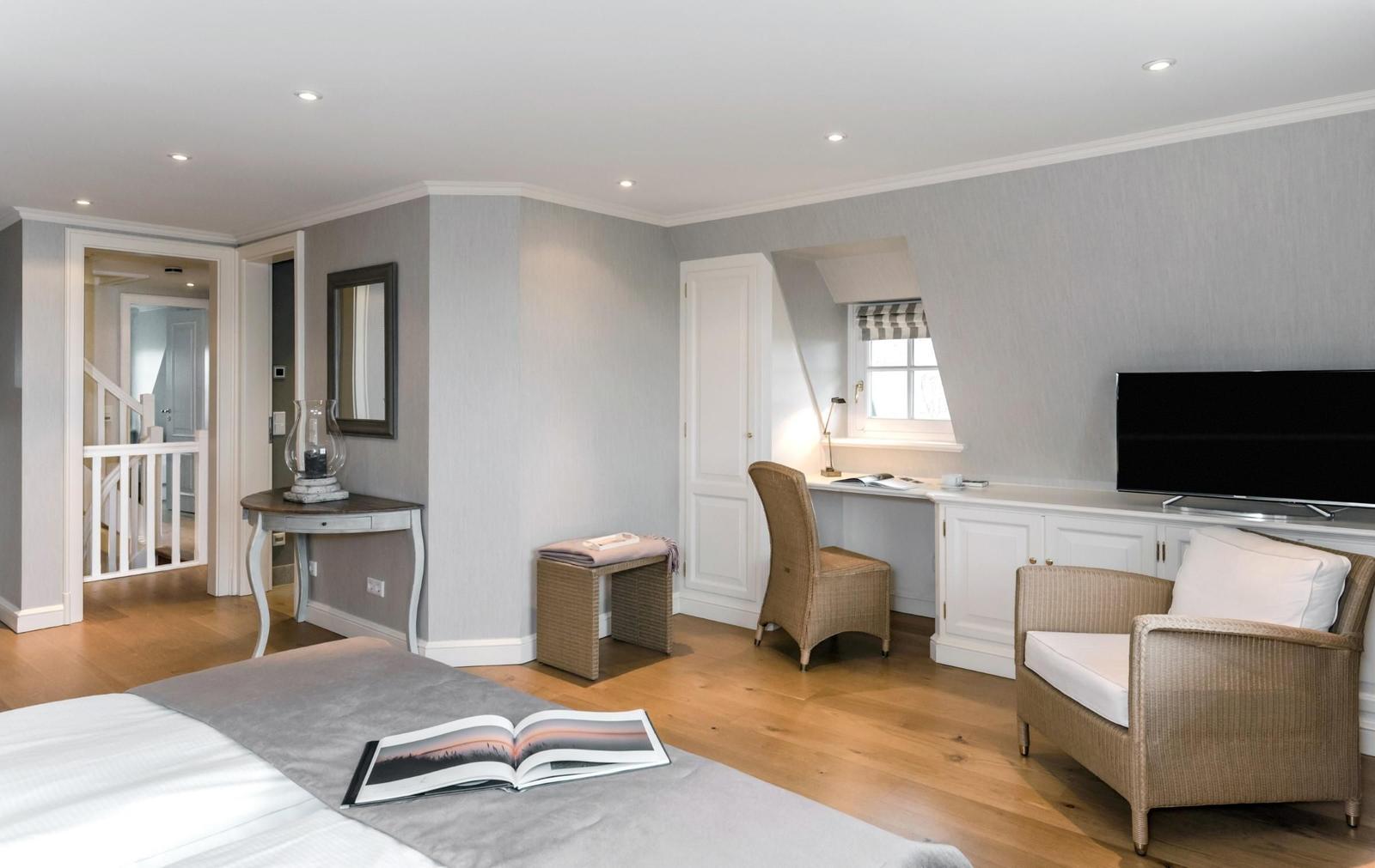 Schlafzimmer mit Bett, Schreibtisch, Sessel und Fernseher. Holzboden und Fenster mit Jalousie.