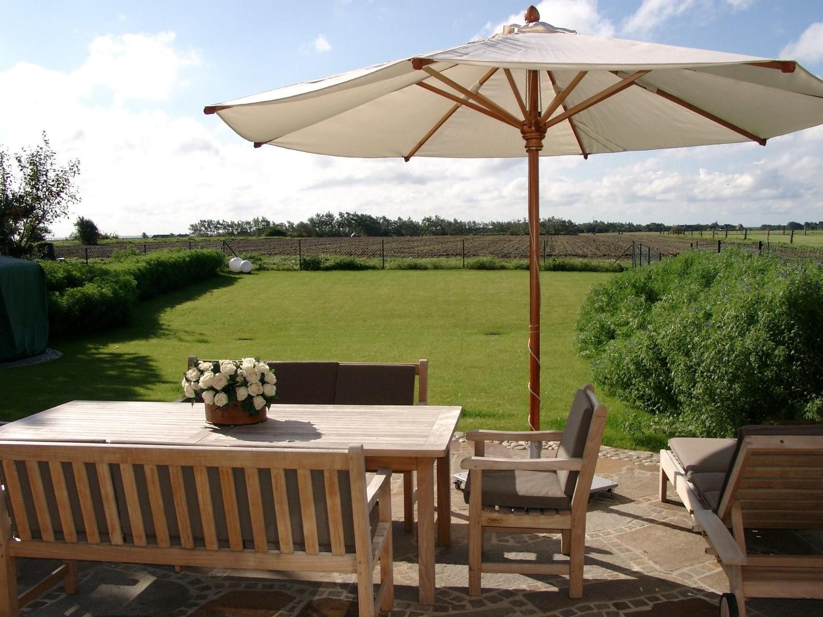 Terrasse mit Tisch, Stühlen und Sonnenschirm. Grünfläche und Felder im Hintergrund.