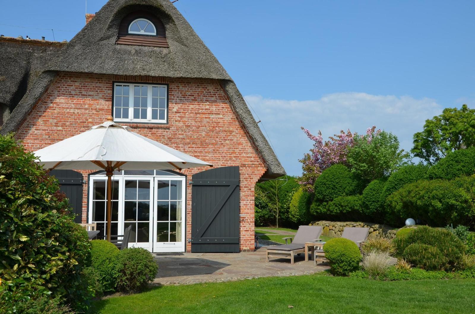 Rotes Backsteingebäude mit Strohdach, Terrasse mit Sonnenschirm und Liegestühlen im Garten.