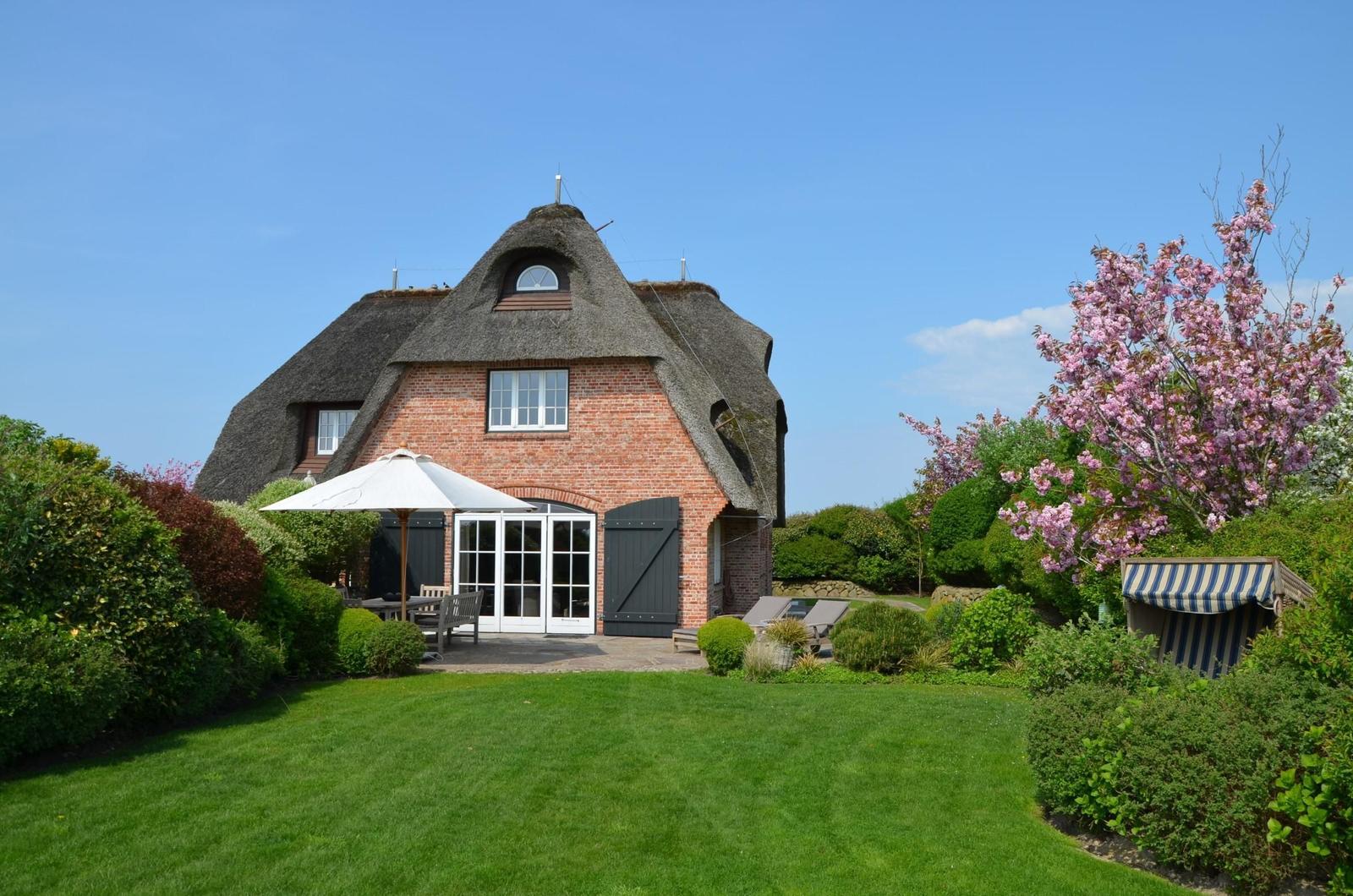 Haus mit Strohdach, Garten, Sonnenliegen und blühender Baum.