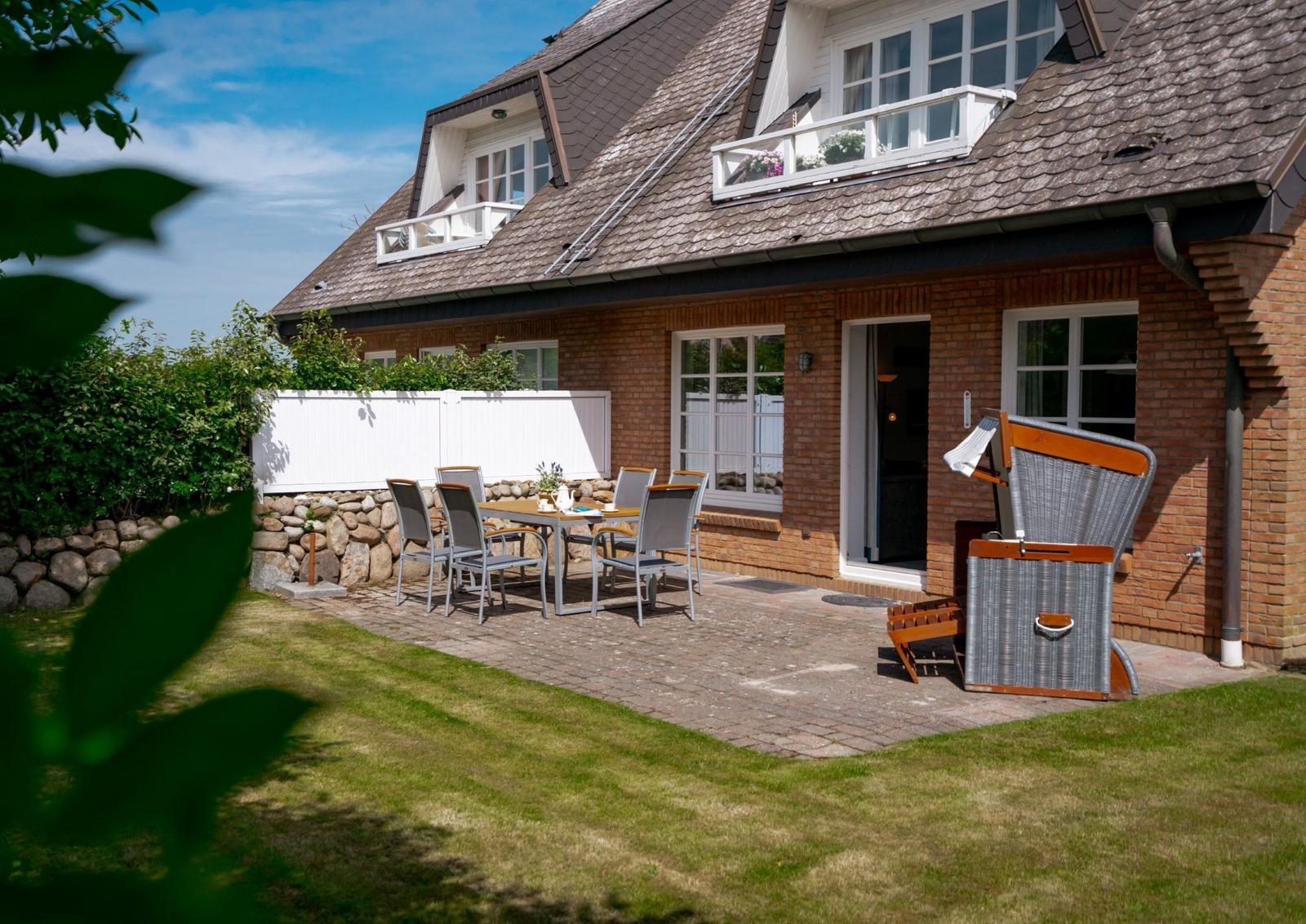 Haus mit Terrasse, Garten und Liegestuhl. Holztisch mit Stühlen vor dem Eingang.