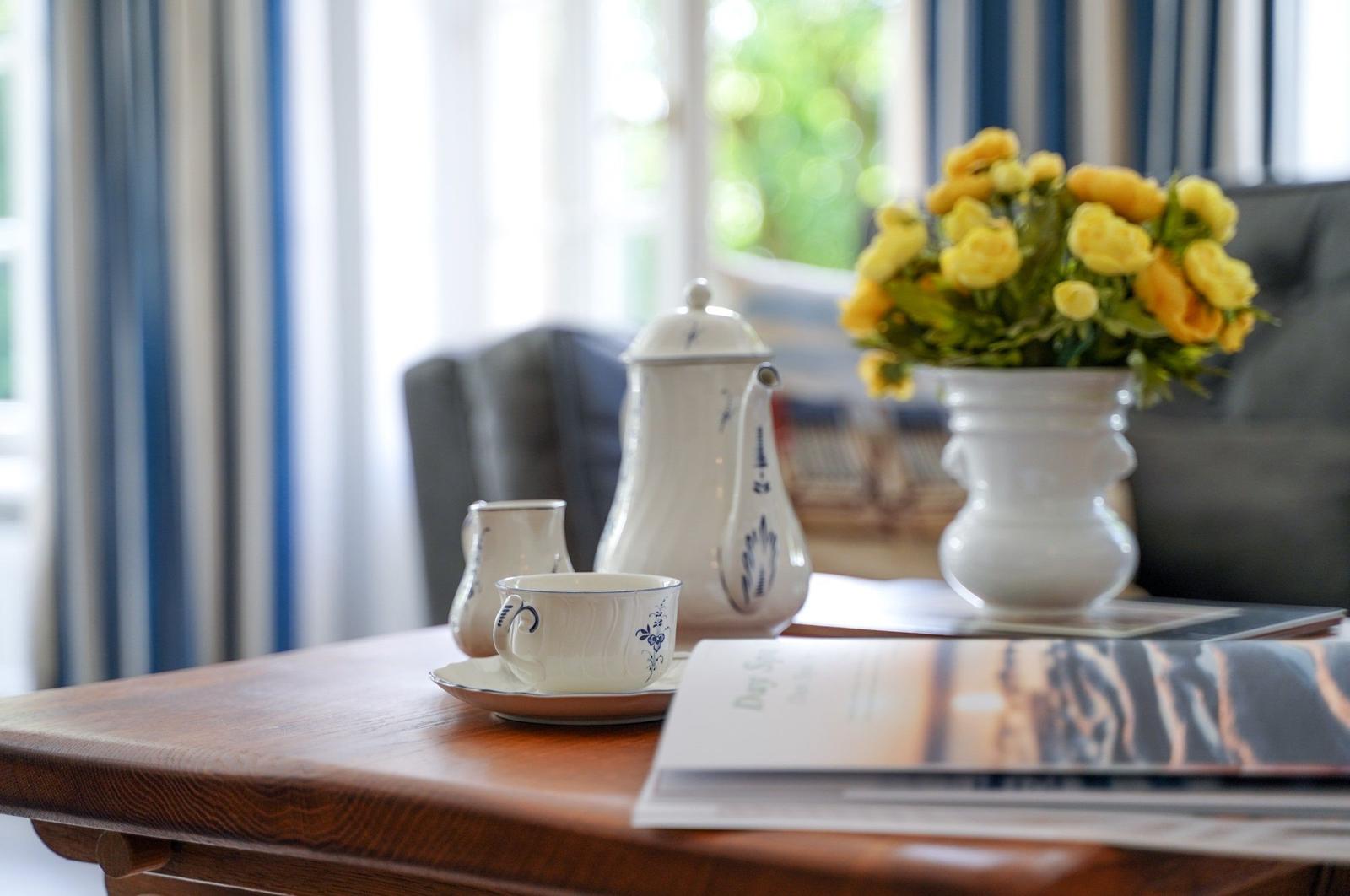 Holztisch mit Teeservice, Blumen und Zeitschriften vor Fenster mit Vorhängen.