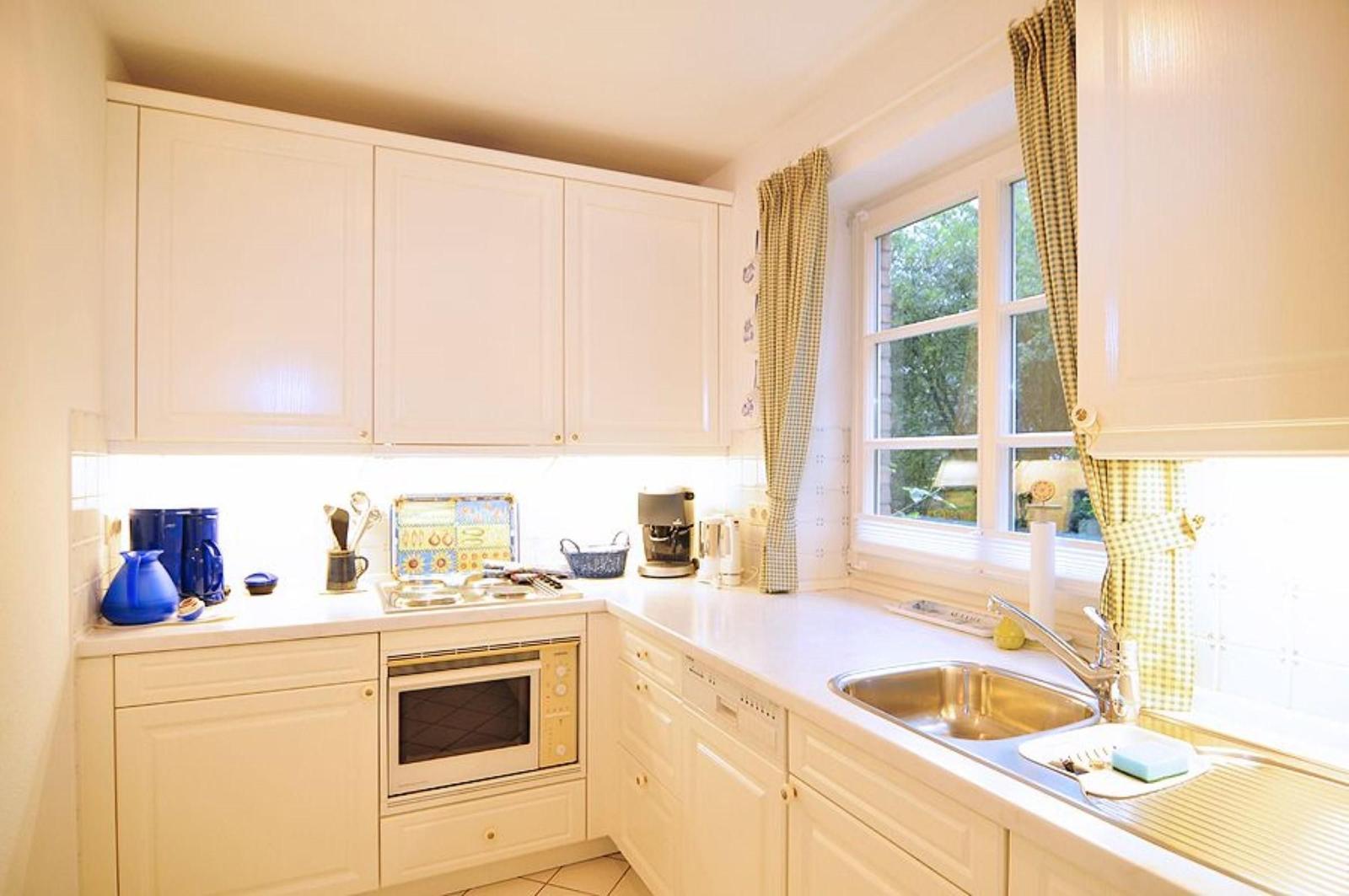 Küche mit weißen Schränken, Spüle und Fenster mit Vorhängen.