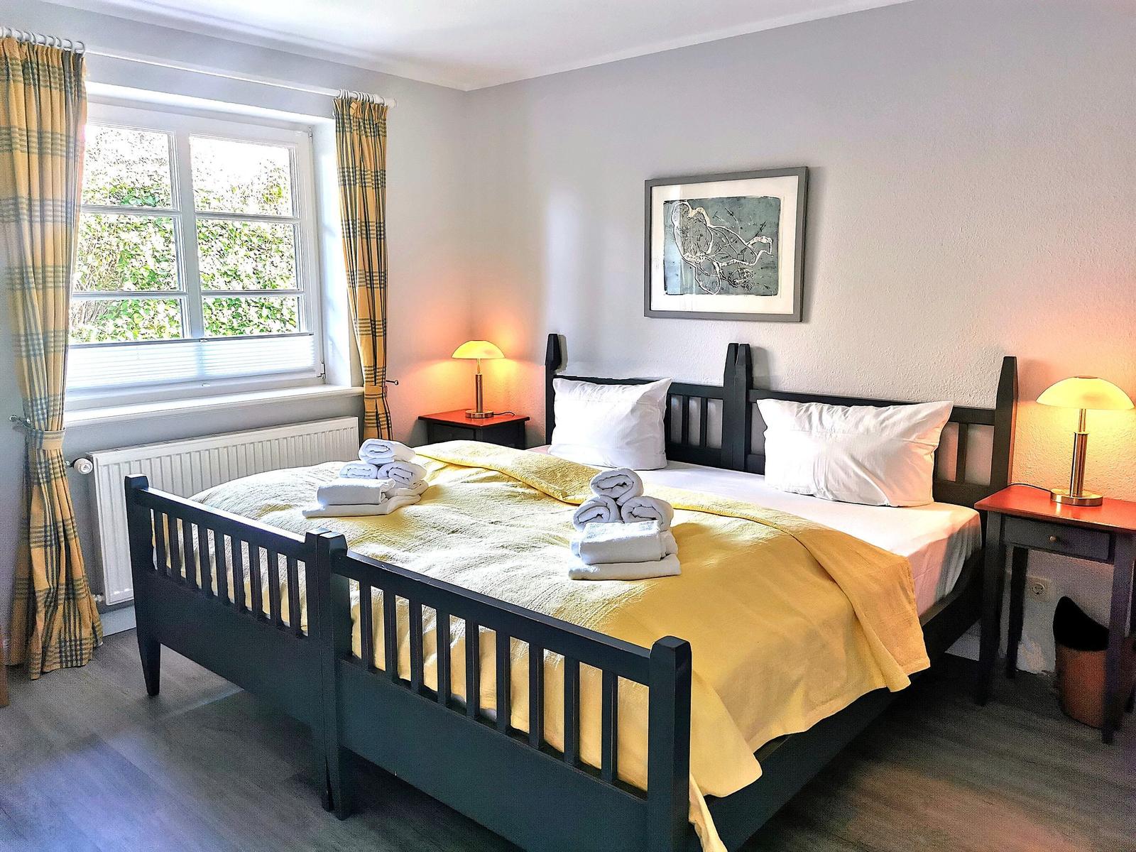 Schlafzimmer mit Bett, Nachttischen und Fenster. Gelbe Bettwäsche und karierte Vorhänge.