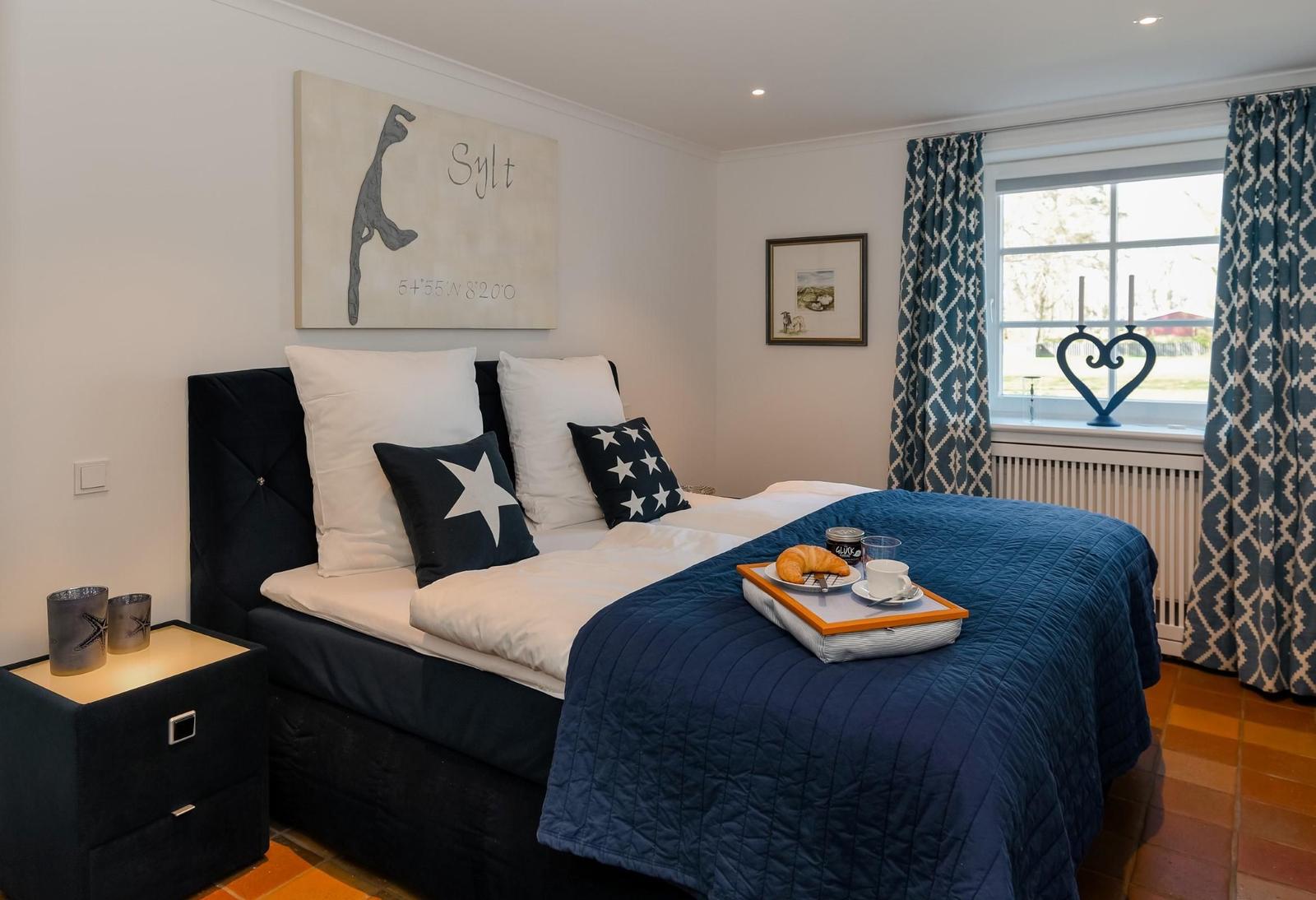 Schlafzimmer mit Bett, Kissen und Frühstückstablett. Fenster mit Vorhängen und Wandbild.