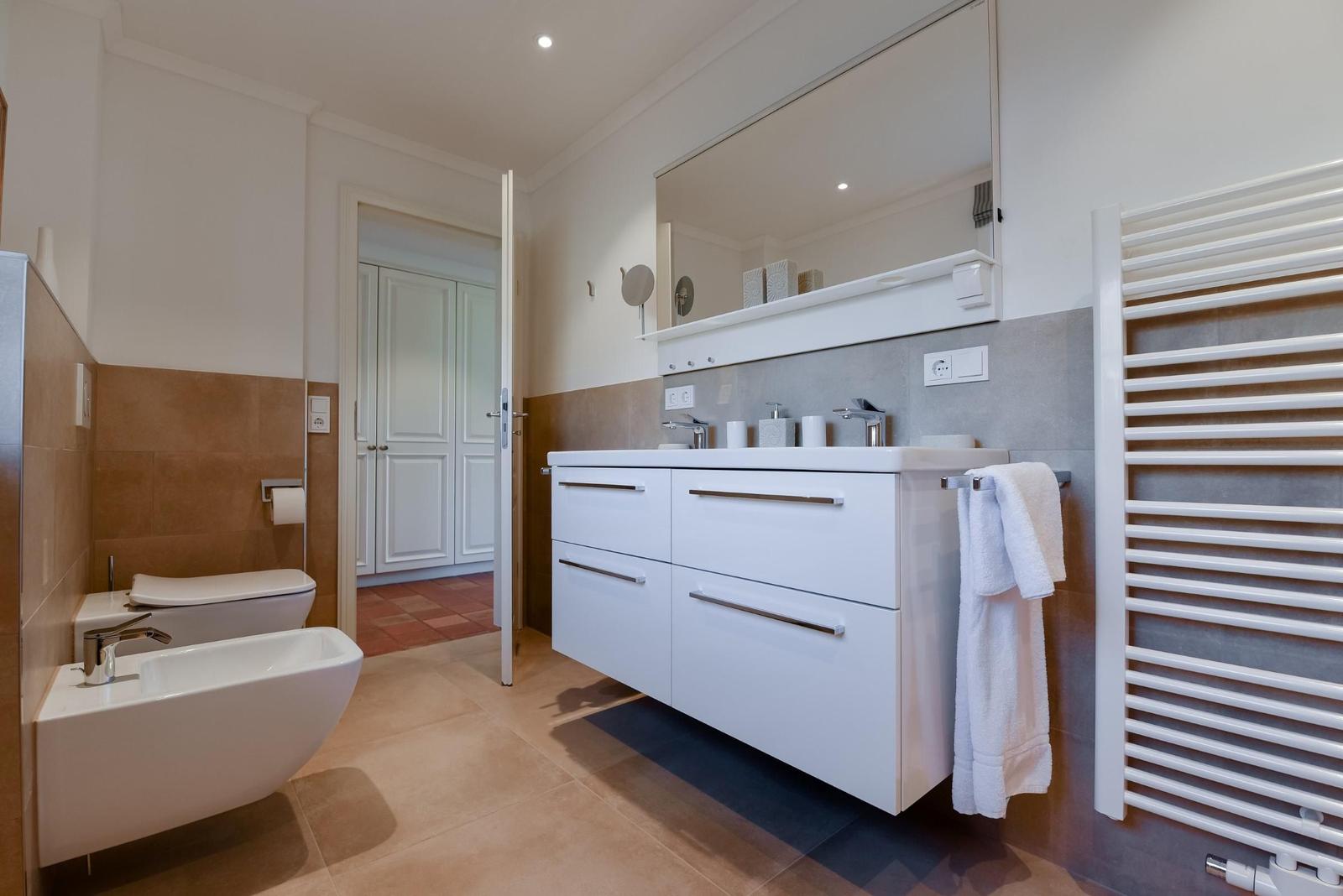 Badezimmer mit Waschtisch, Spiegel und Toilette. Warme Fliesen und weißer Schrank.