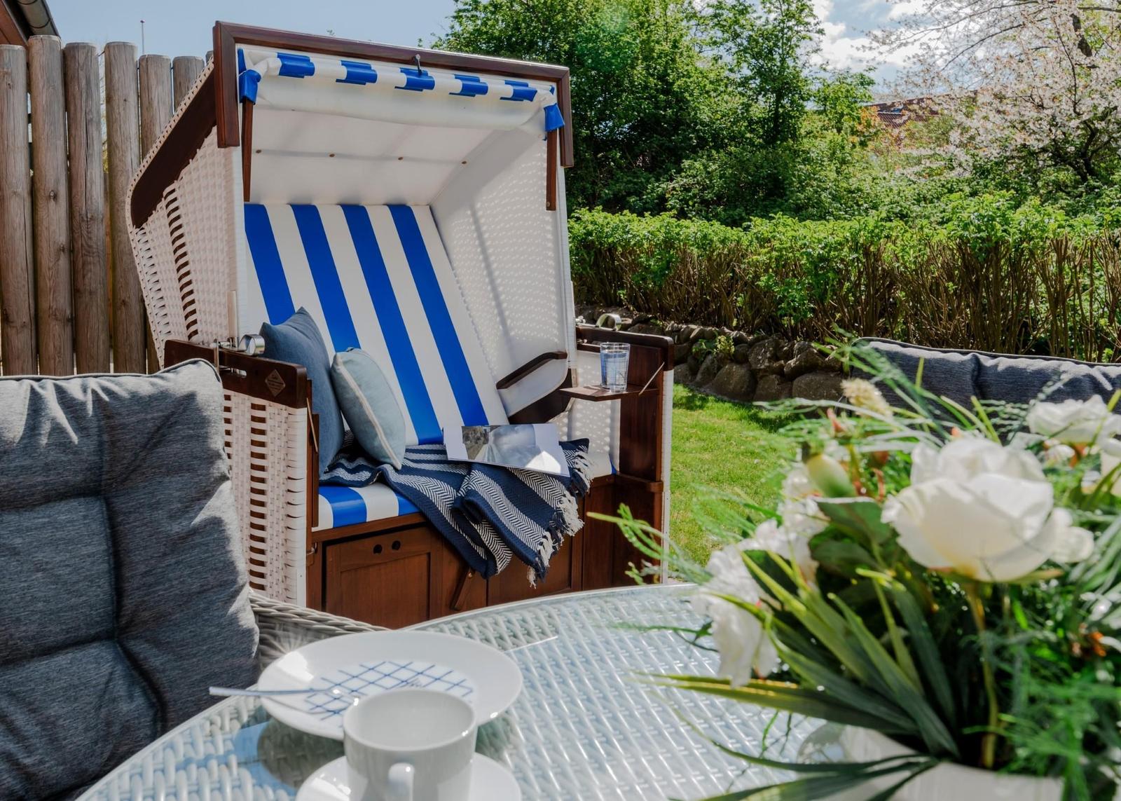 Terrasse mit Strandkorb, Tisch und Blumen. Hintergrund: grüner Garten.