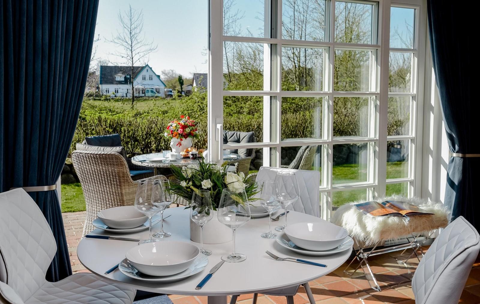 Runder Tisch mit Geschirr und Gläsern vor großem Fenster mit Blick auf Garten.