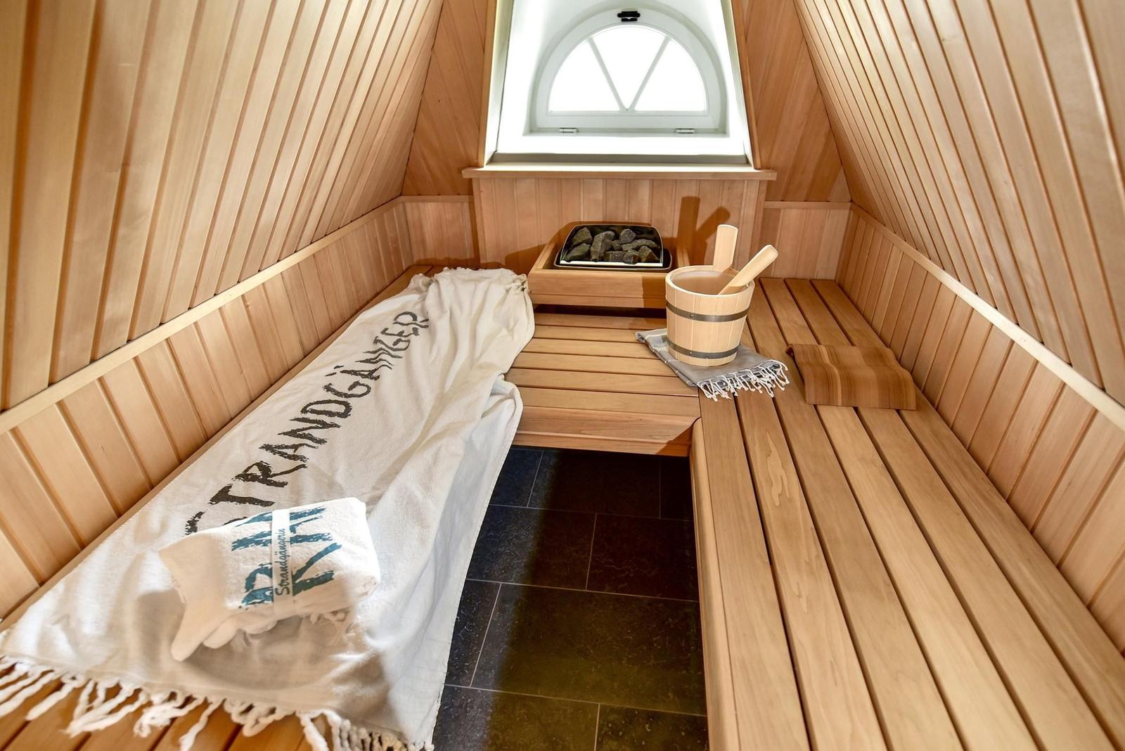 Sauna mit Holzbank, Steinmulde und Eimer. Fenster mit Bogenform. Decke und Wände aus Holz.