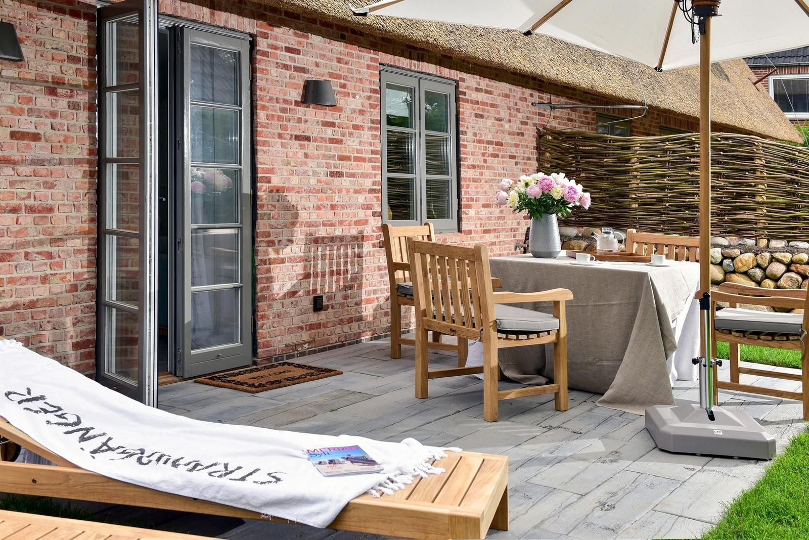 Terrasse mit Holzgartenmöbeln, Tisch mit Blumen und Sonnenschirm. Steinmauer und Ziegelwand im Hintergrund.