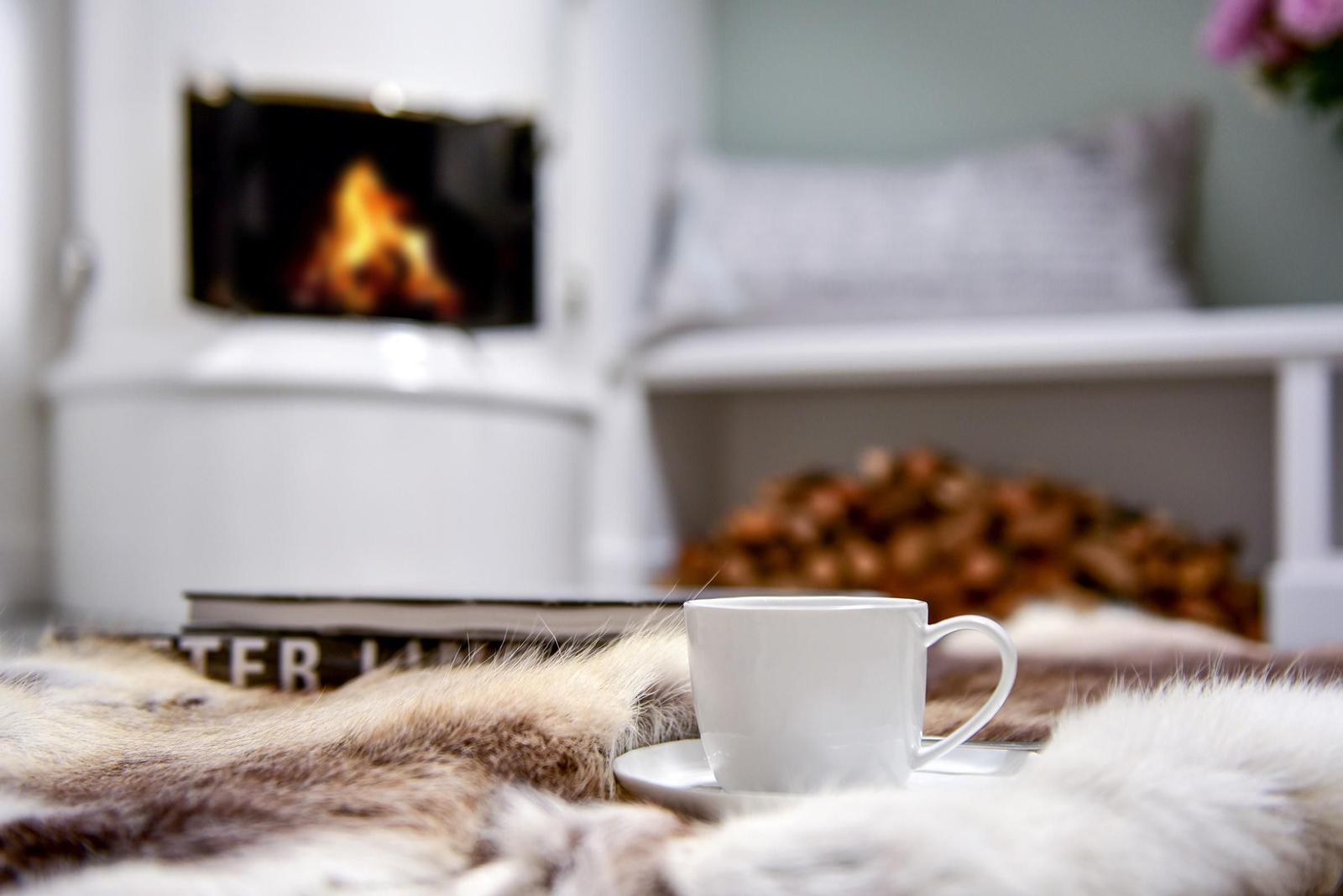 Eine weiße Tasse steht auf einem Fell, vor einem Kamin mit brennendem Feuer.