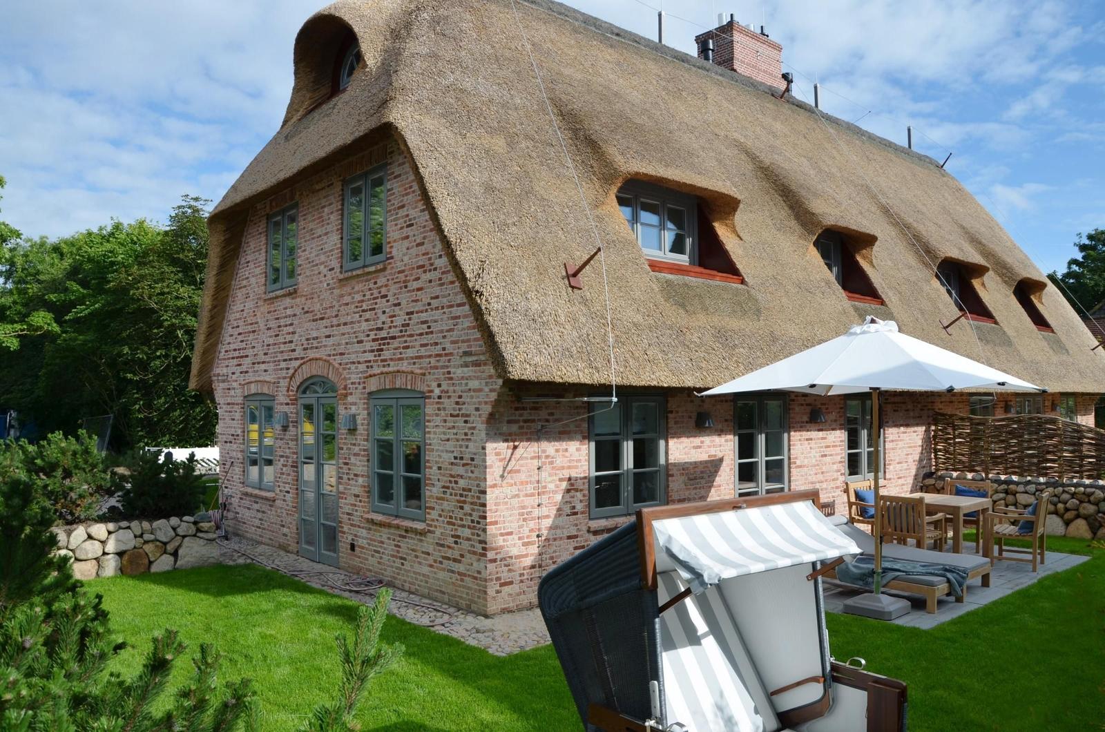 Haus mit Strohdach, Terrasse mit Sonnenschirm und Liegestühlen im Garten.