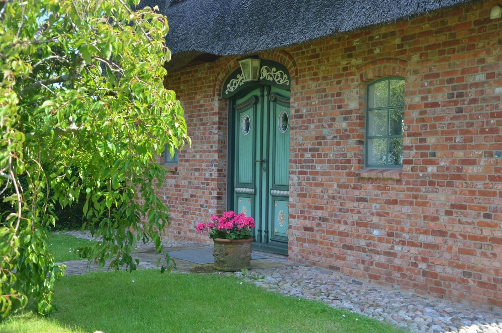 Ein roter Ziegelbau mit grüner Tür und Dach aus Stroh. Vor dem Eingang steht ein Blumentopf.