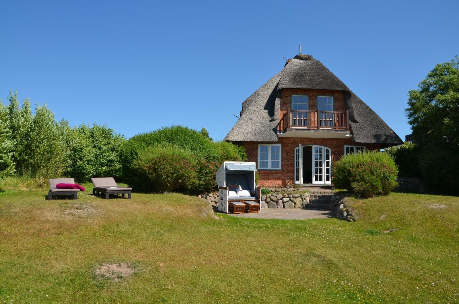 Rotes Backsteingebäude mit Strohdach, Terrasse und Garten mit Liegen.