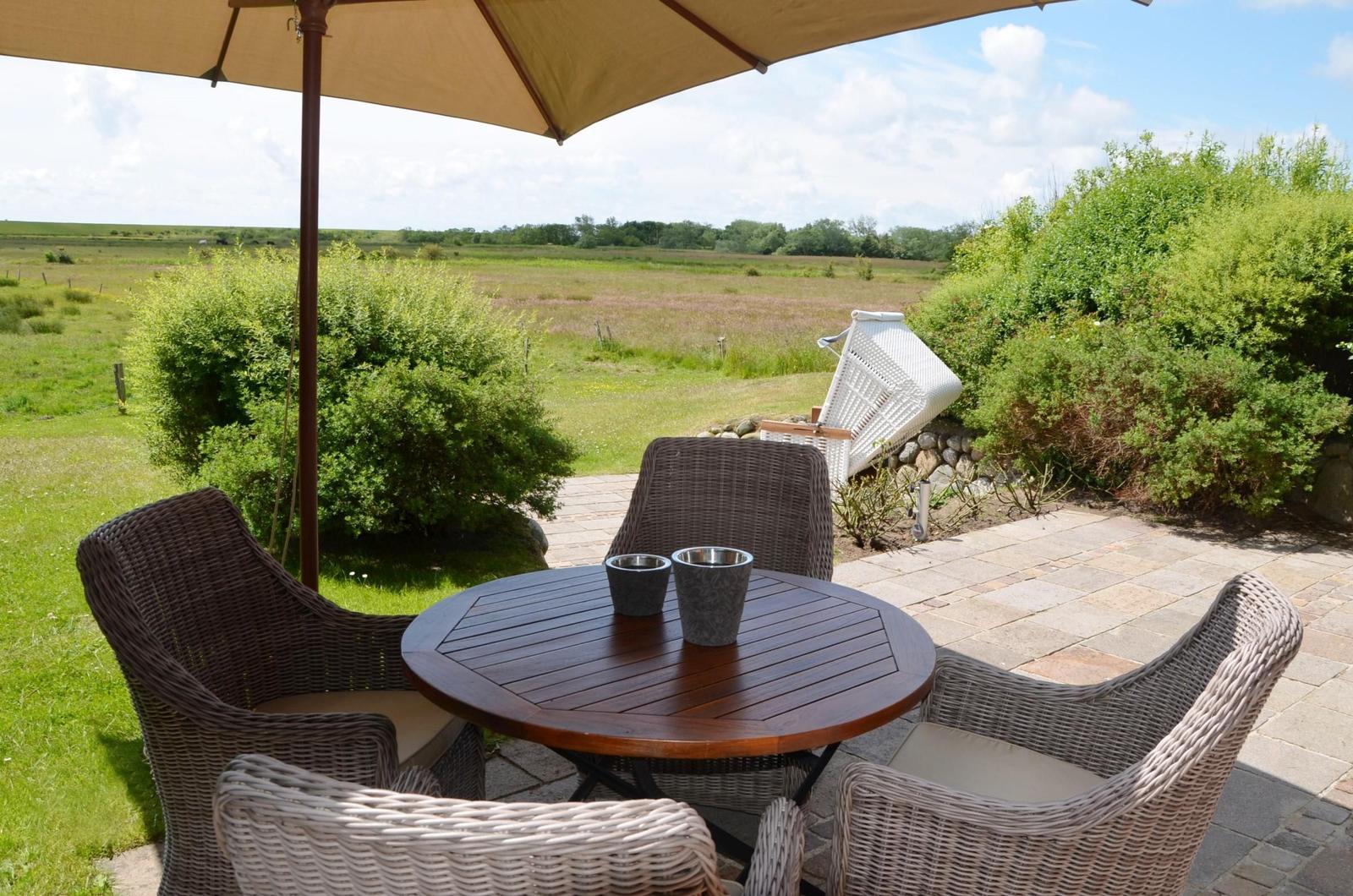 Terrasse mit Tisch, Stühlen und Sonnenschirm. Blick auf grüne Wiesen.