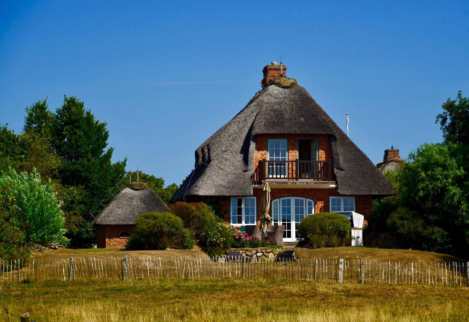 Haus mit Strohdach, Balkon und Garten. Umgeben von Bäumen und Gras.