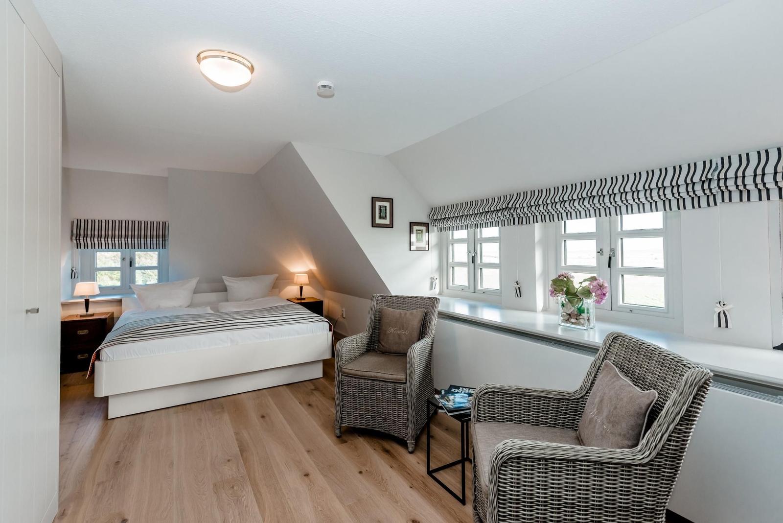 Bedroom with sloped ceiling, bed, two chairs, and windows with roller blinds.