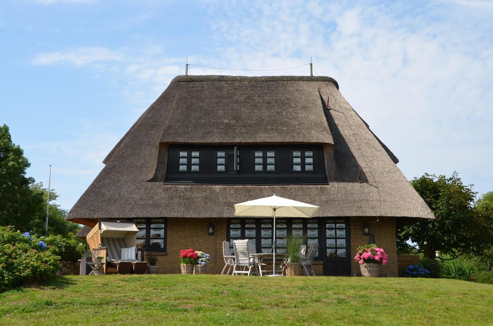House with thatched roof, terrace, and garden. White sunshade and flower pots.
