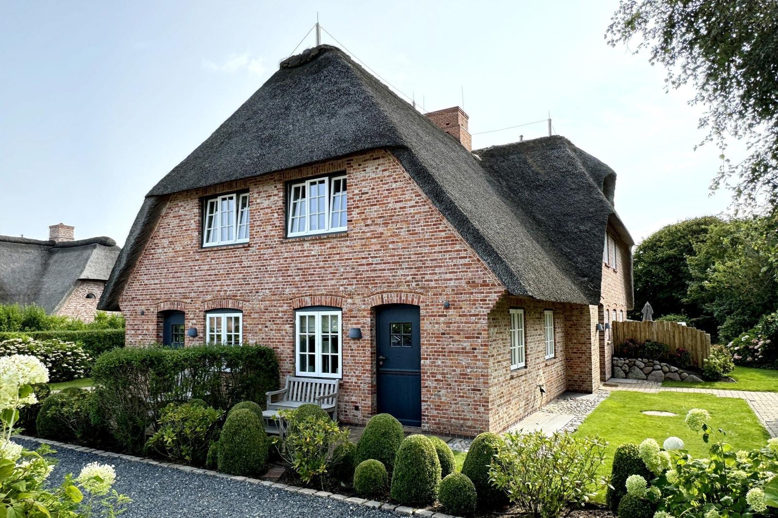 Haus mit Strohdach, roten Ziegeln und blauer Tür. Garten mit Gras und Sträuchern.