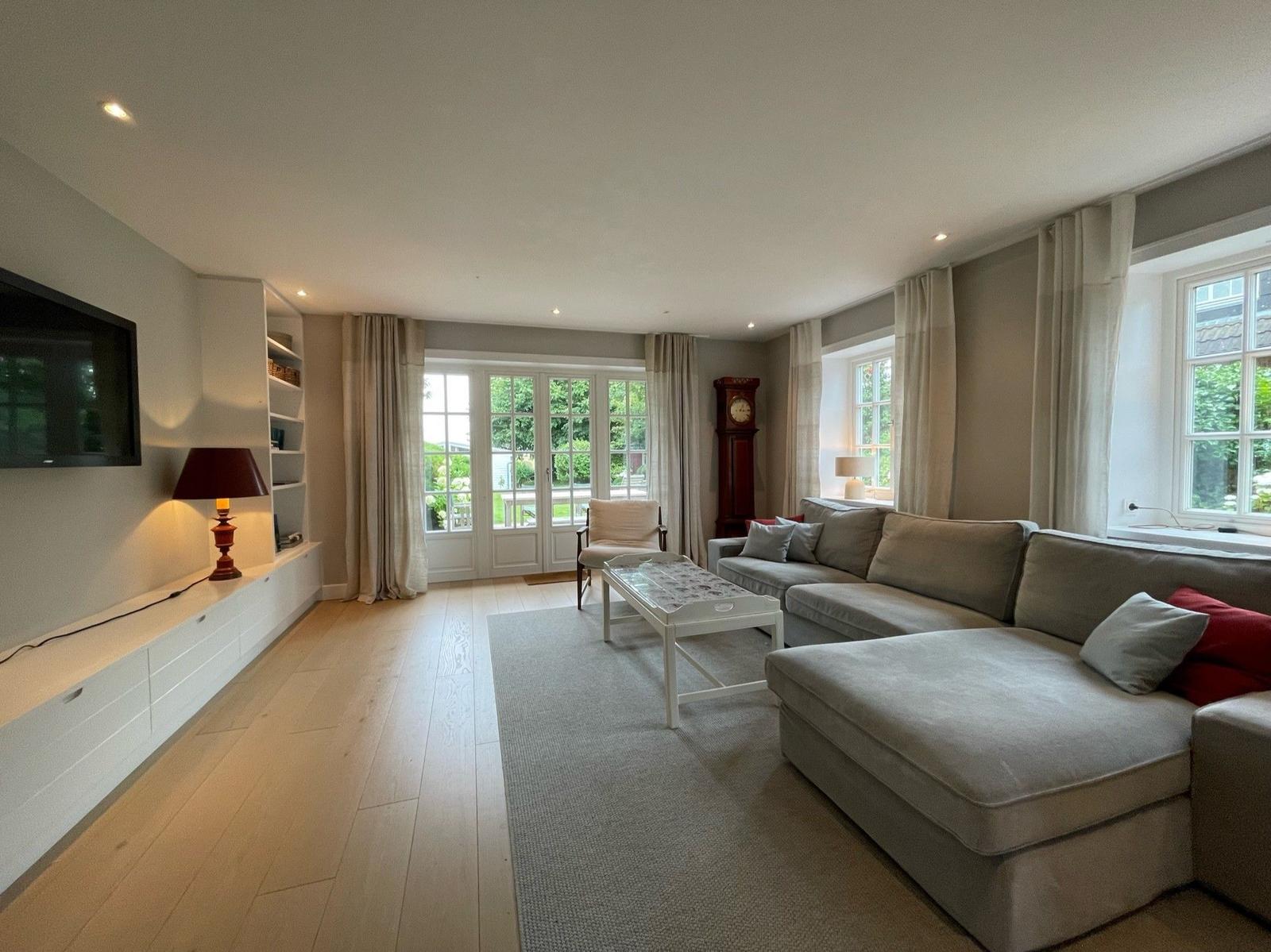 Wohnzimmer mit Sofa, Couchtisch und TV-Wand. Große Fenster und helle Holzböden.