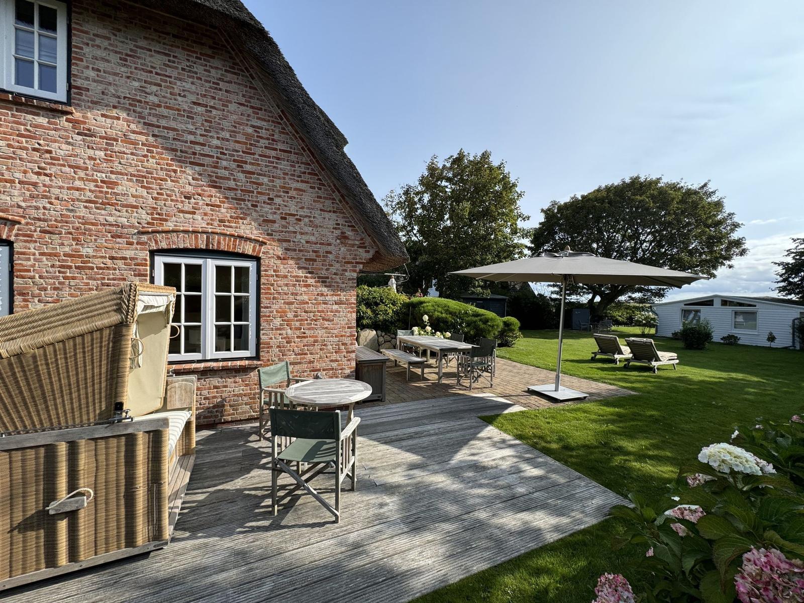 Holzterrasse mit Tischen, Stühlen und Sonnenschirm neben einem Backsteinhaus mit Dach.