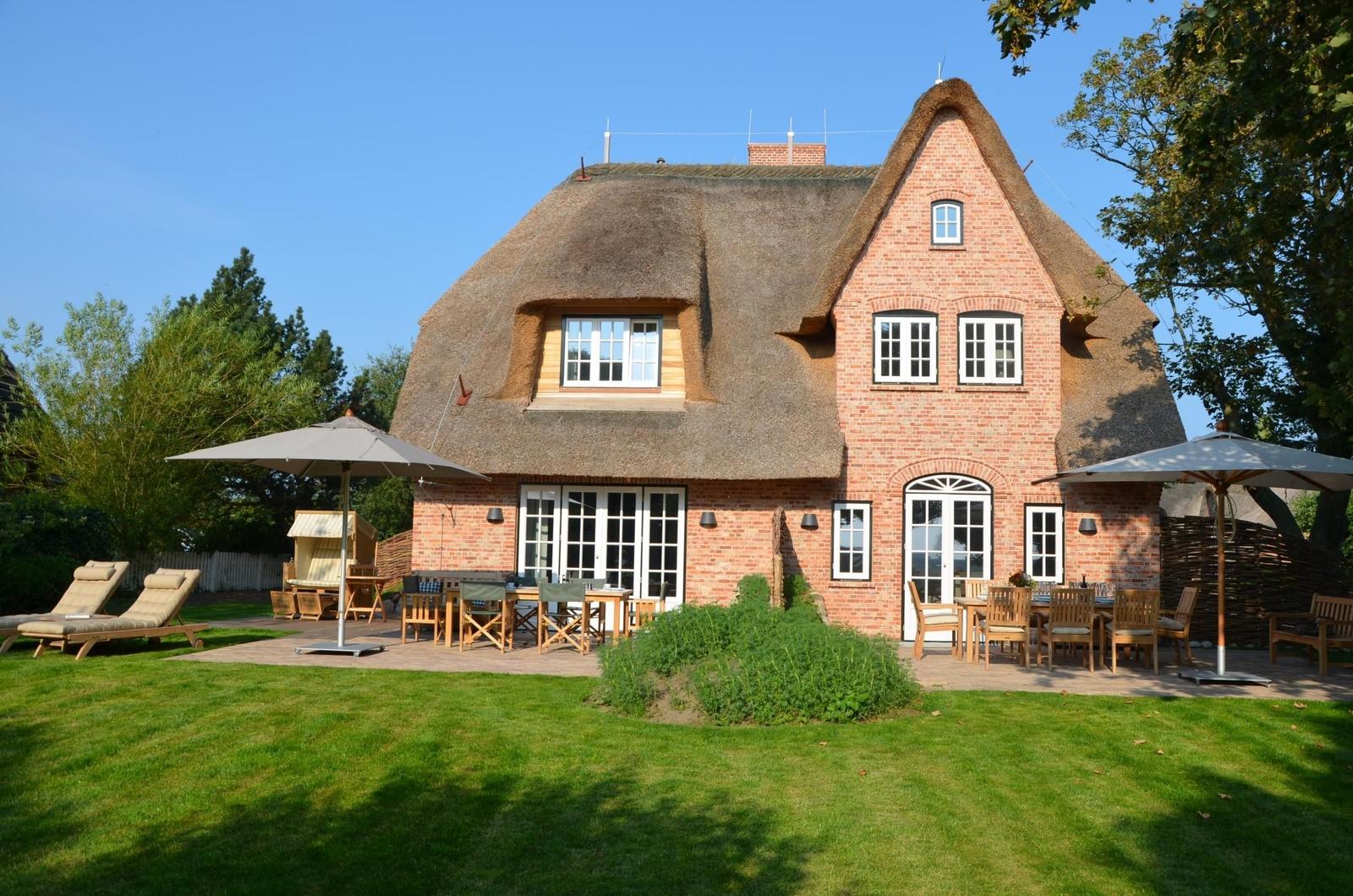 Haus mit Strohdach, Terrasse und Garten mit Liegestühlen und Tischen.