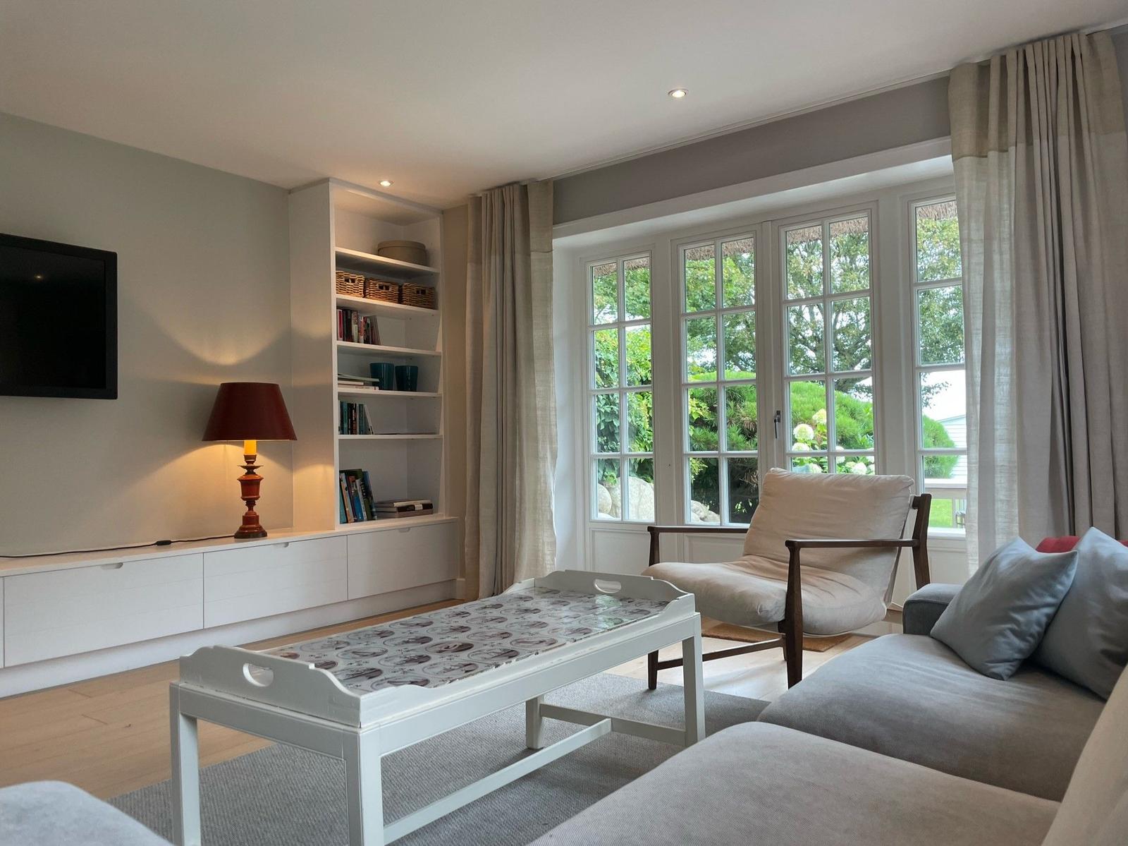 Wohnzimmer mit Sofa, Sessel, Fernseher und Bücherregal. Große Fenster mit Blick auf Garten.