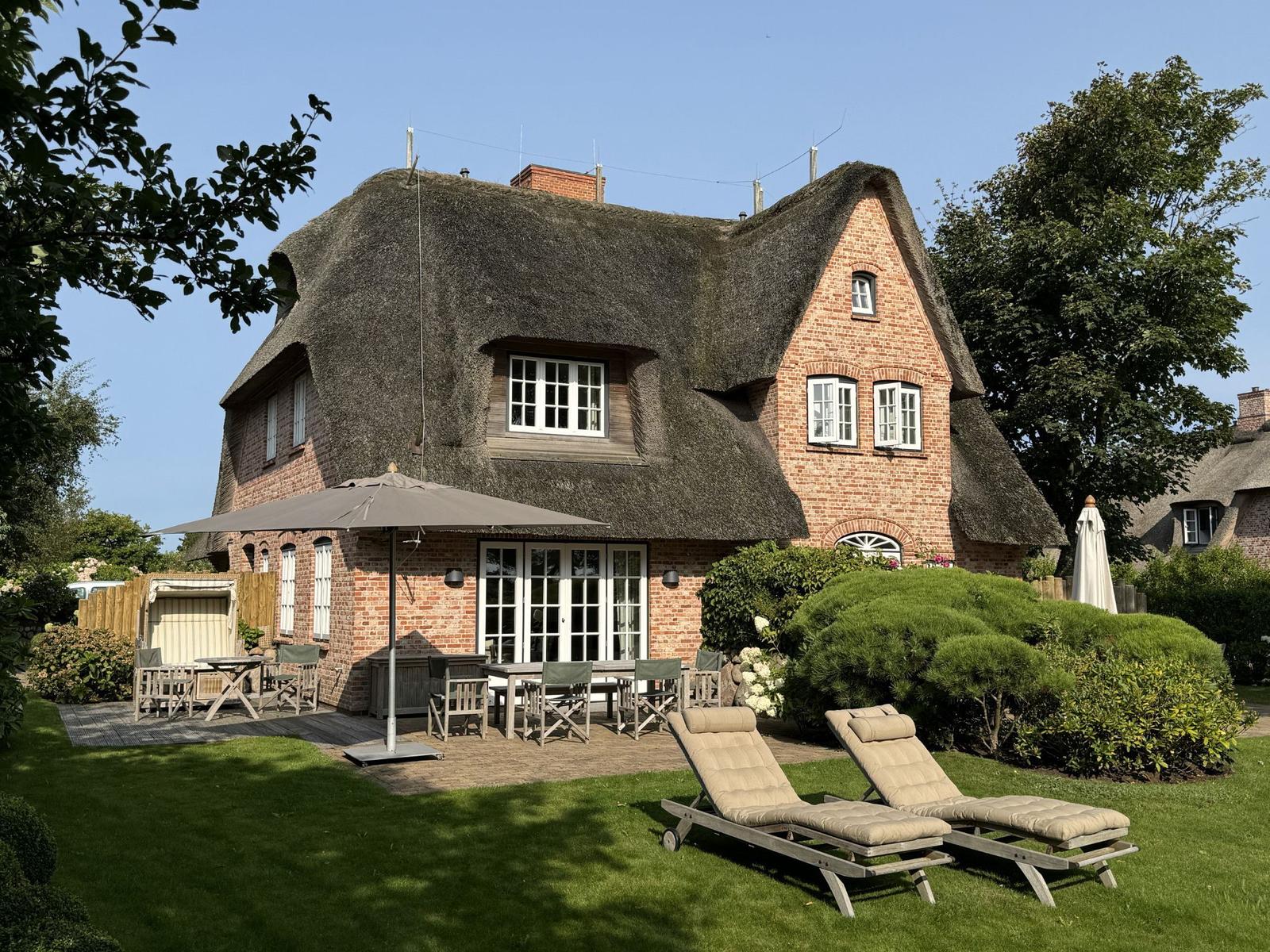 Dorfhaus mit Strohdach, Terrasse und Gartenliegen.